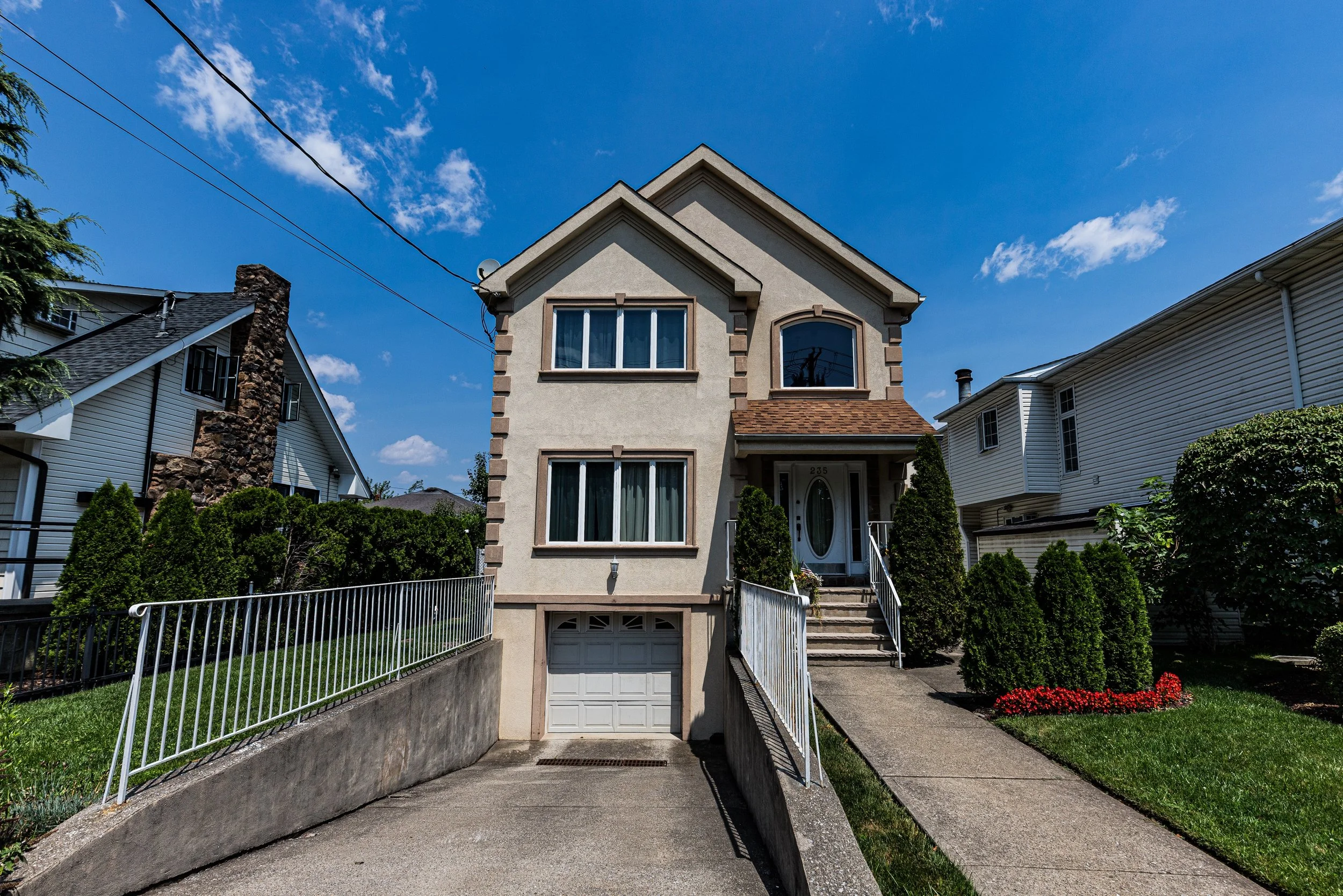 A two-story beige house with a garage at street level, front steps leading to the door, and well-maintained garden with bushes and flowers, under a blue sky with some clouds.