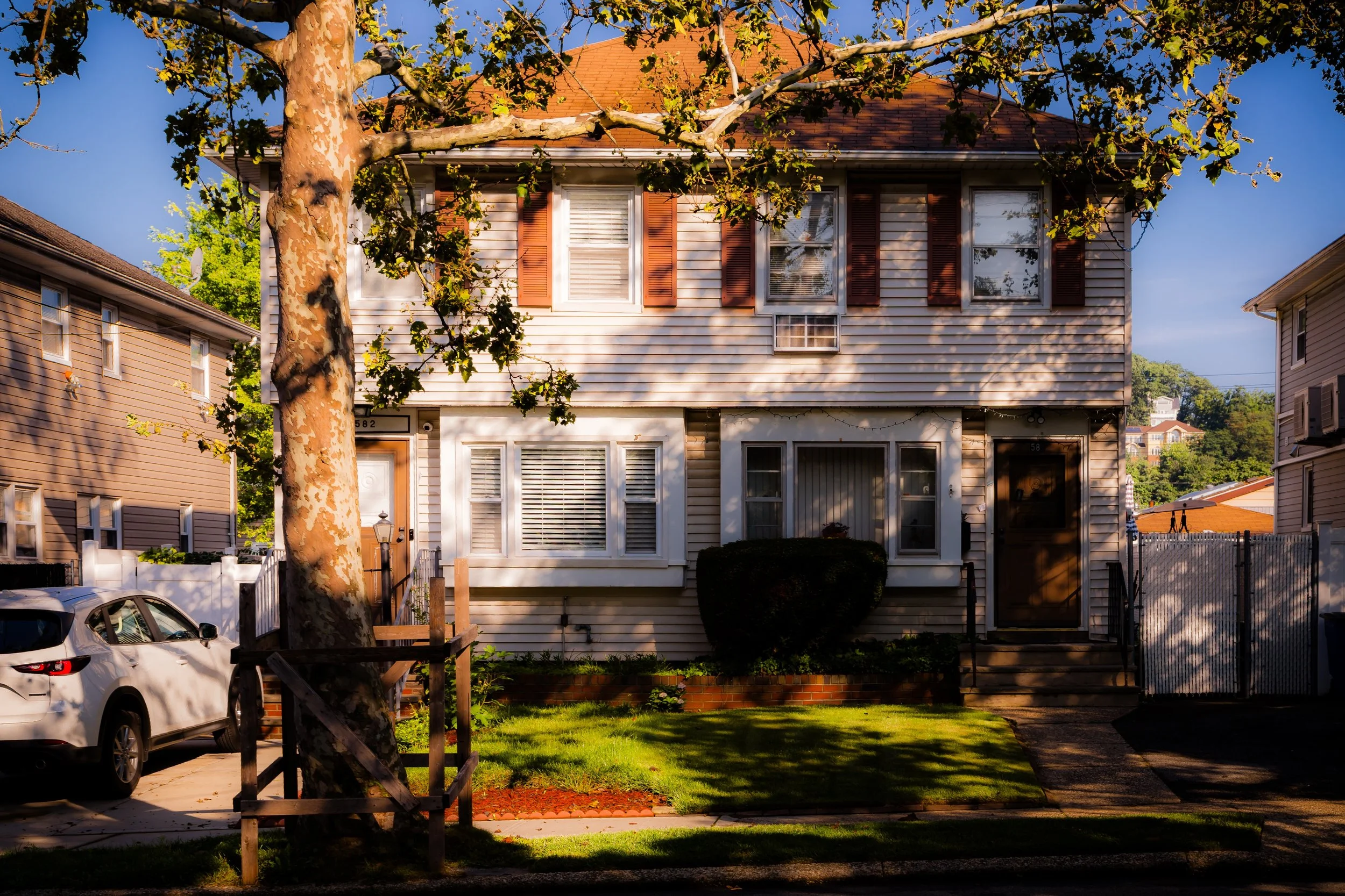 A two-story house with beige siding, brown shutters, and a brown front door. There is a small front lawn with greenery and a trimmed shrub near the house. A large tree with peeling bark and green leaves stands in front, casting shadows. A white car is parked in the driveway on the left side of the image, and neighboring houses are visible on both sides. The sky is clear and blue, indicating a sunny day.