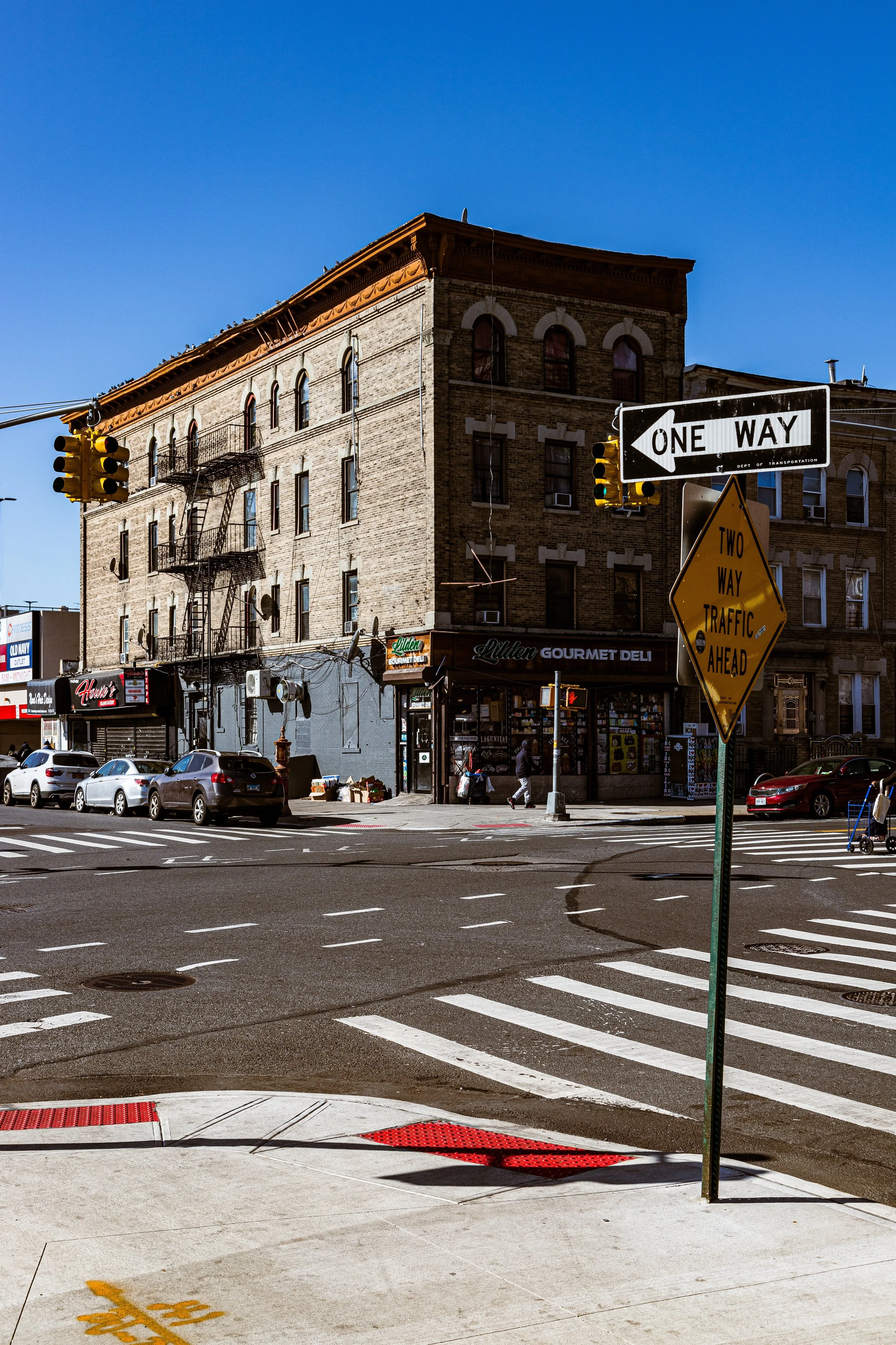 A city street corner with multi-story brick buildings, parked cars, traffic lights, and street signs indicating one-way and two-way traffic.