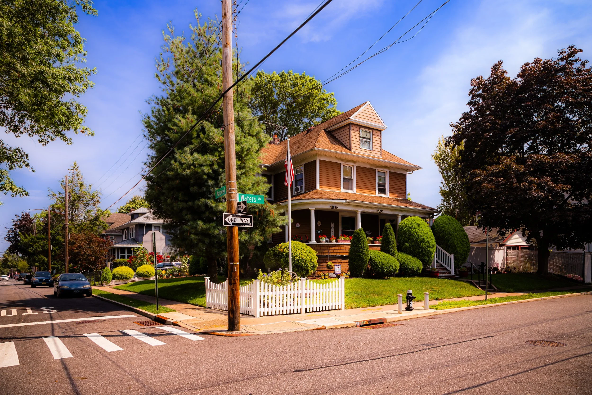 A corner house on Watters Avenue with a well-maintained front yard, white picket fence, and greenery. The house has a brown exterior, multiple windows, and a porch decorated with potted plants. There are trees and nearby houses visible, and street signs for Watters Avenue and a one-way street on the corner.