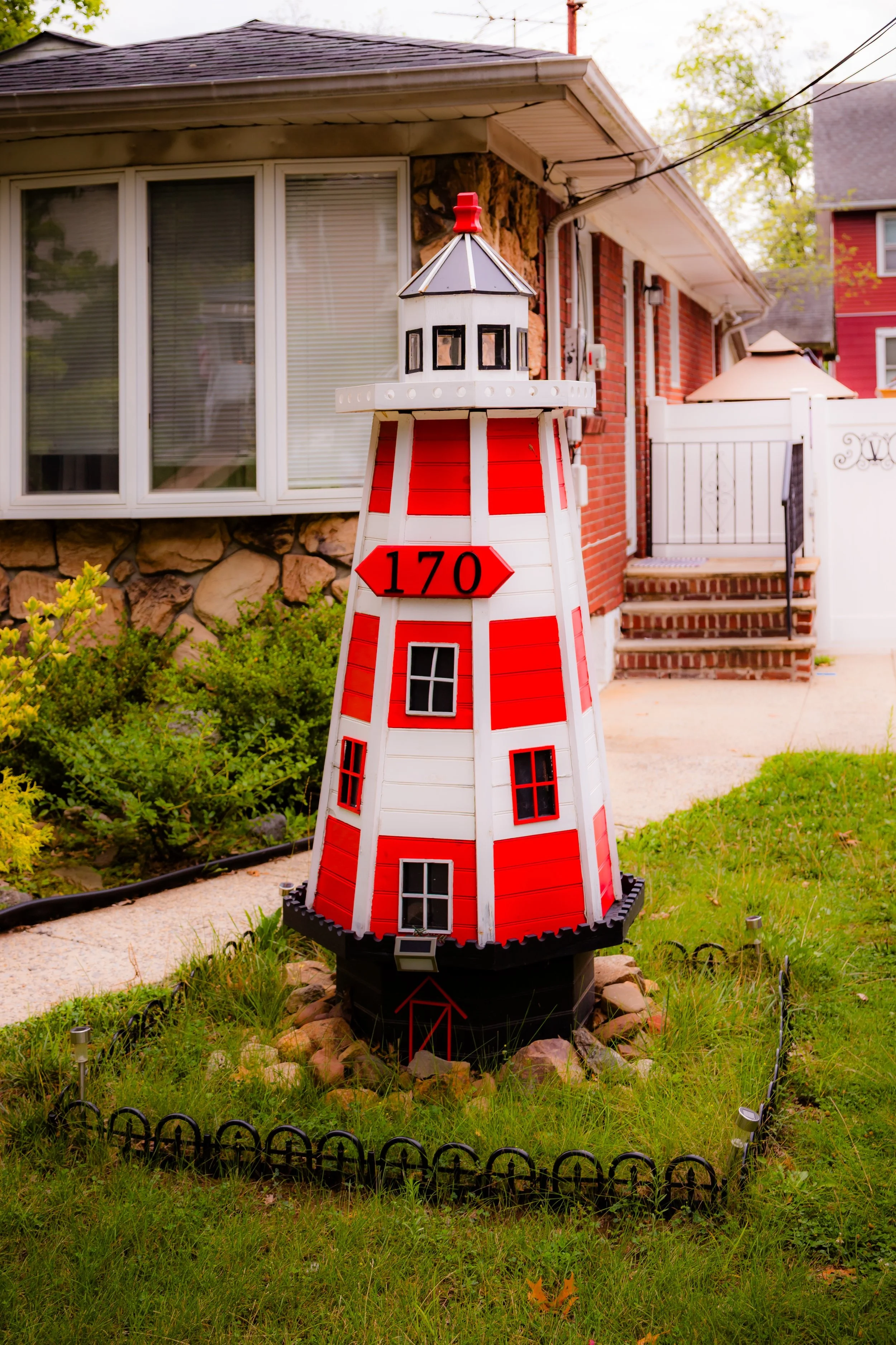 Miniature red and white lighthouse decoration in a front yard, with house and porch in the background.