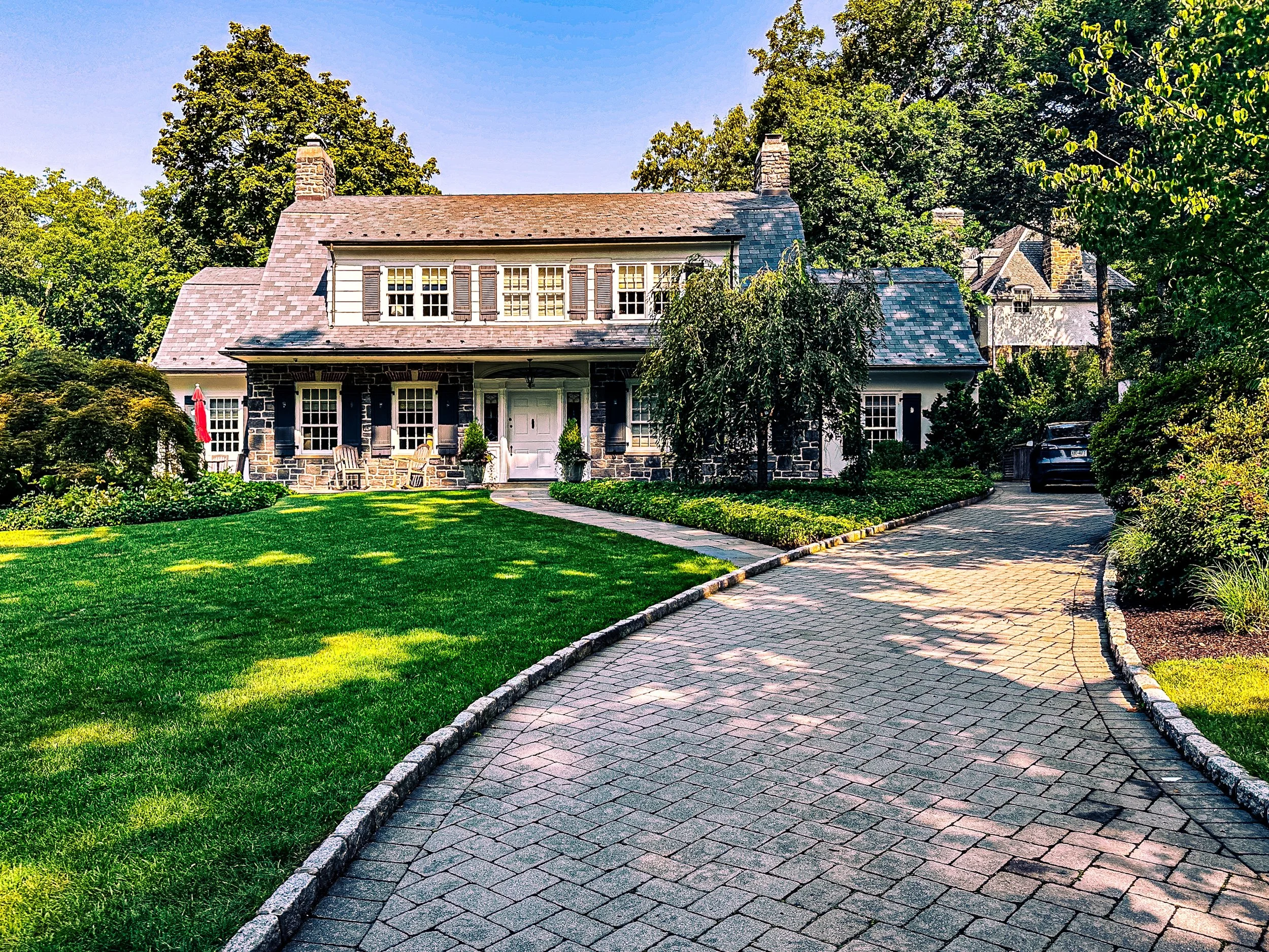 A large house with a stone and shingle exterior, a front porch with seating, surrounded by a well-manicured lawn and trees, with a curved driveway leading to the house and a car parked near the side.