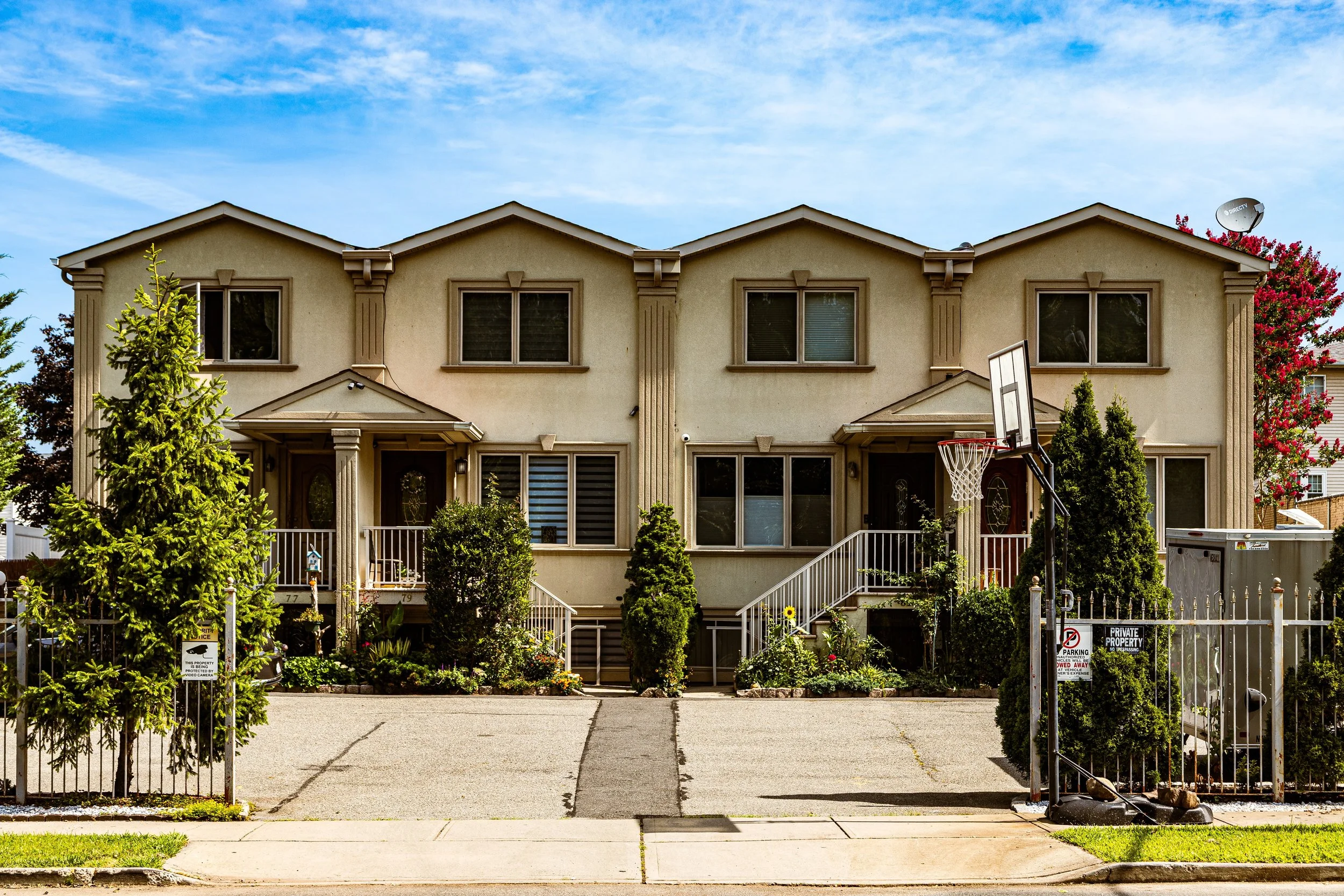 Two-story beige residential building with small front porch and stairs, surrounded by green bushes and trees, with a basketball hoop in the driveway and a satellite dish on the roof.