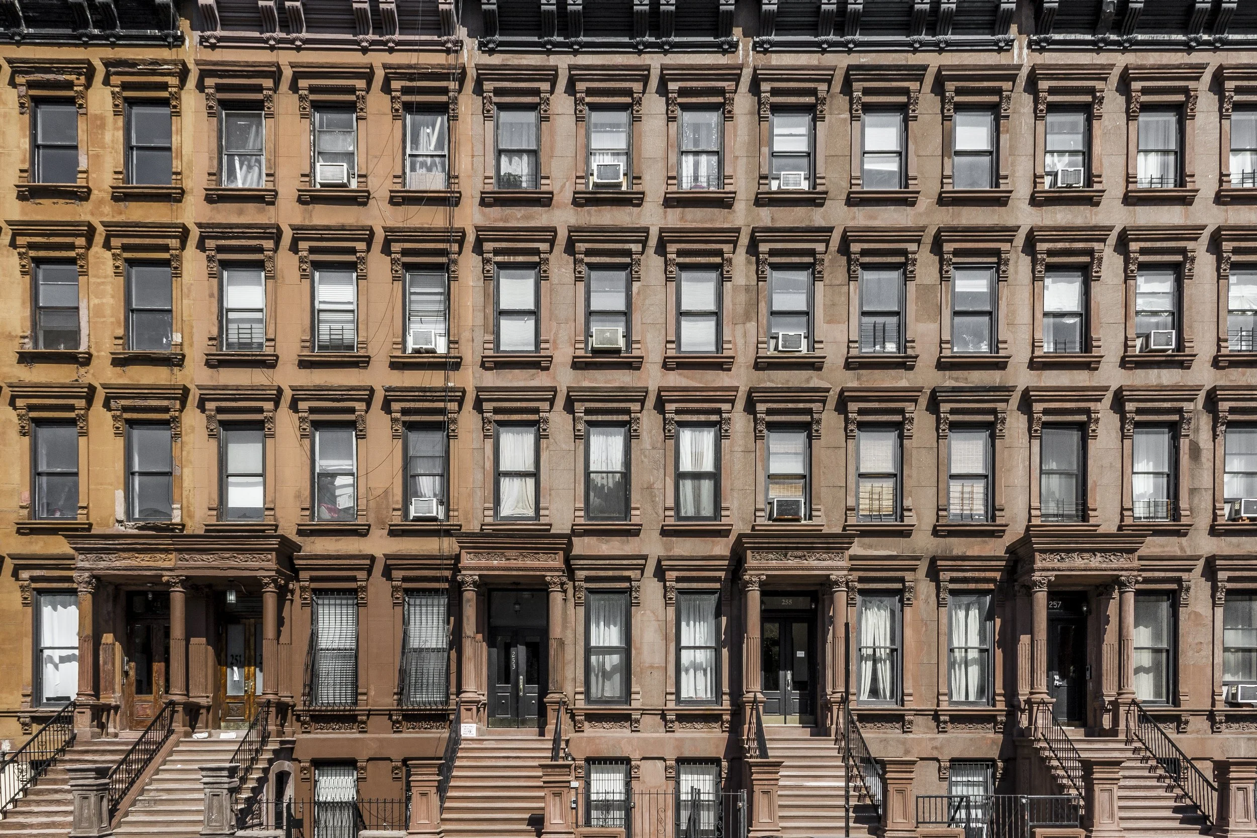 A brownstone apartment building with multiple floors, featuring large windows with black or white window air conditioning units, some with curtains or blinds. The building has front stairs leading to entry doors and decorative architectural details around windows and doors.