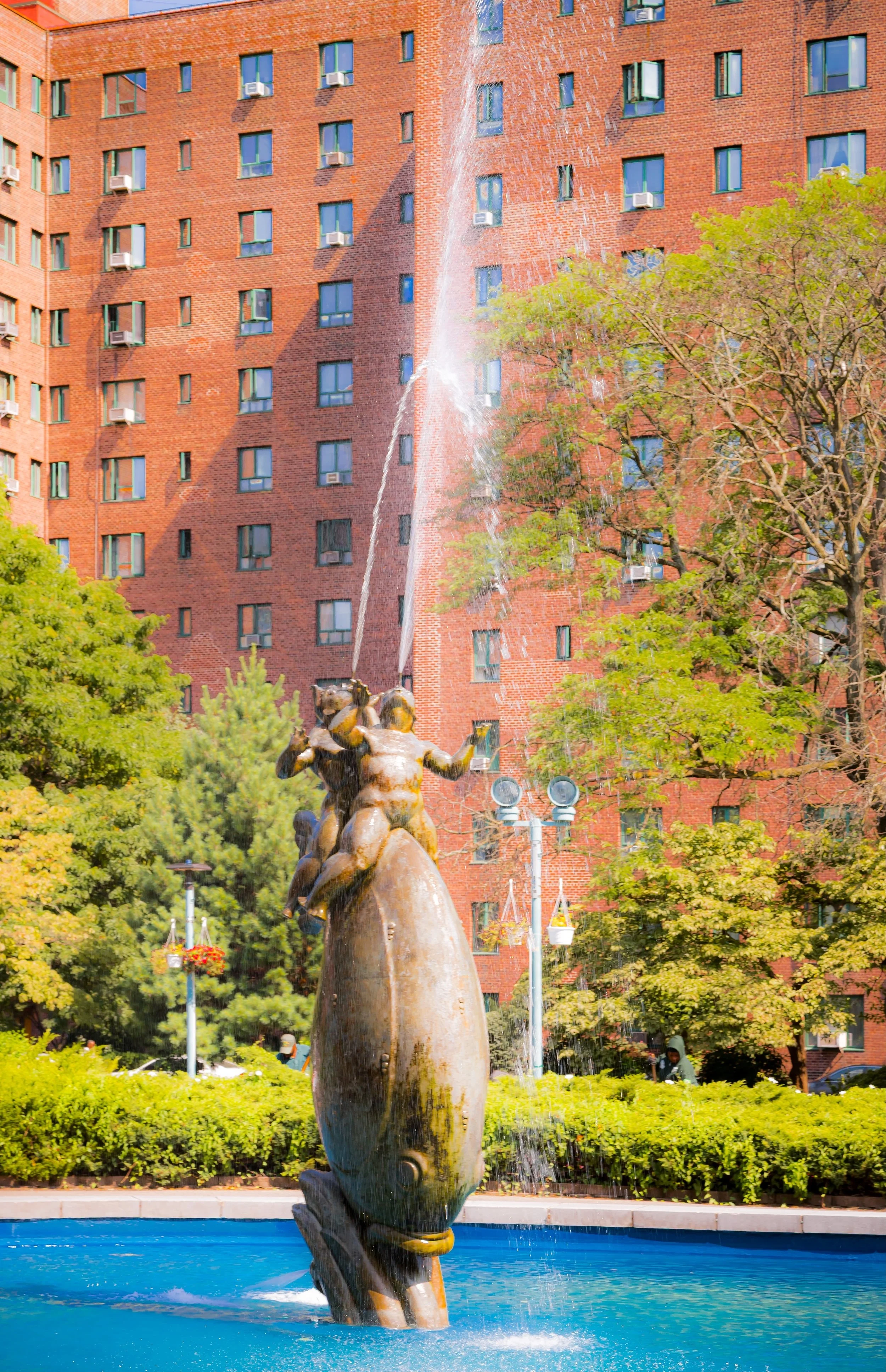 A fountain featuring a sculpture of a fish with water spouting from its mouth, surrounded by greenery and in front of a red brick apartment building.