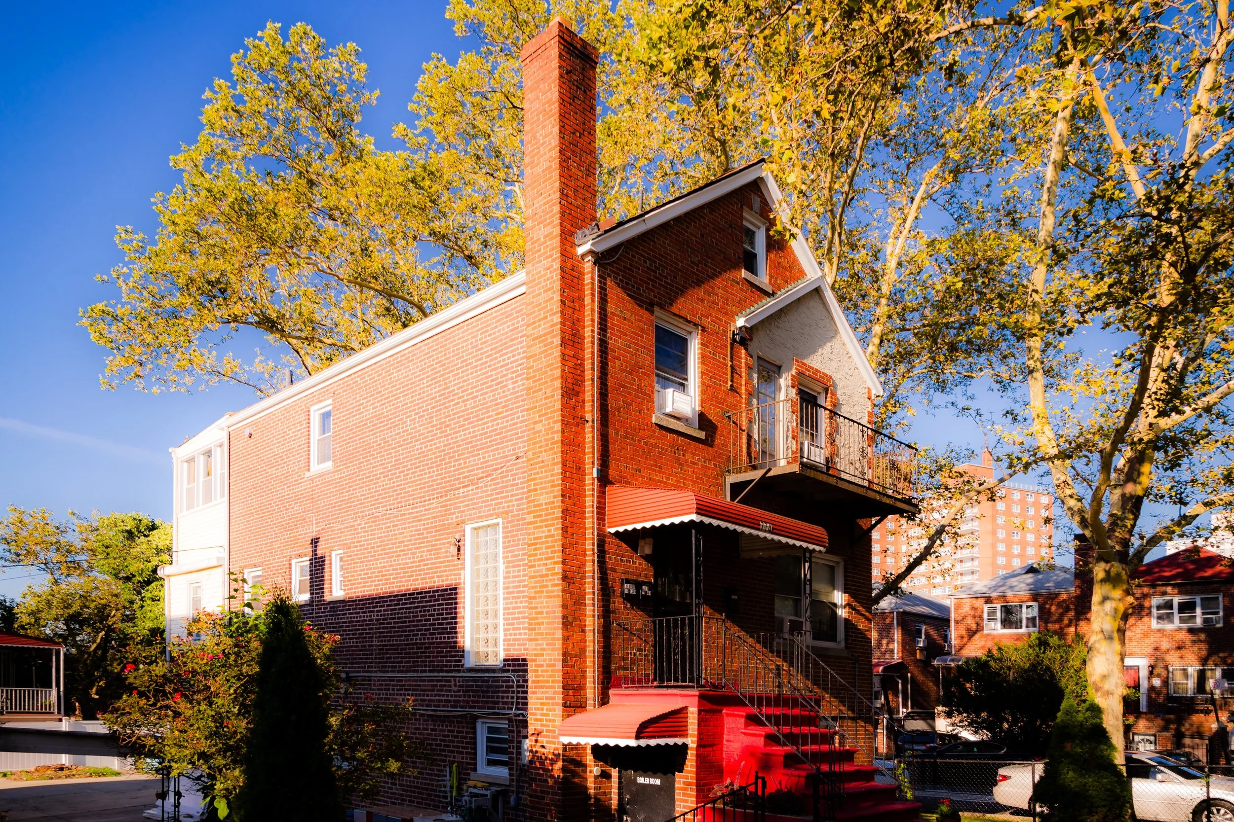 A brick residential building with stairs leading to an upper balcony, surrounded by trees with yellow leaves, and a blue sky in the background.