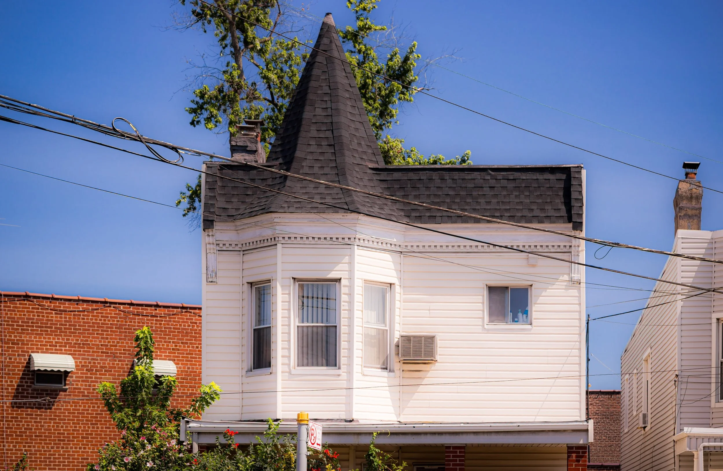 A white, two-story house with a steep, pointed roof and bay windows, surrounded by trees and overhead power lines, against a clear blue sky.