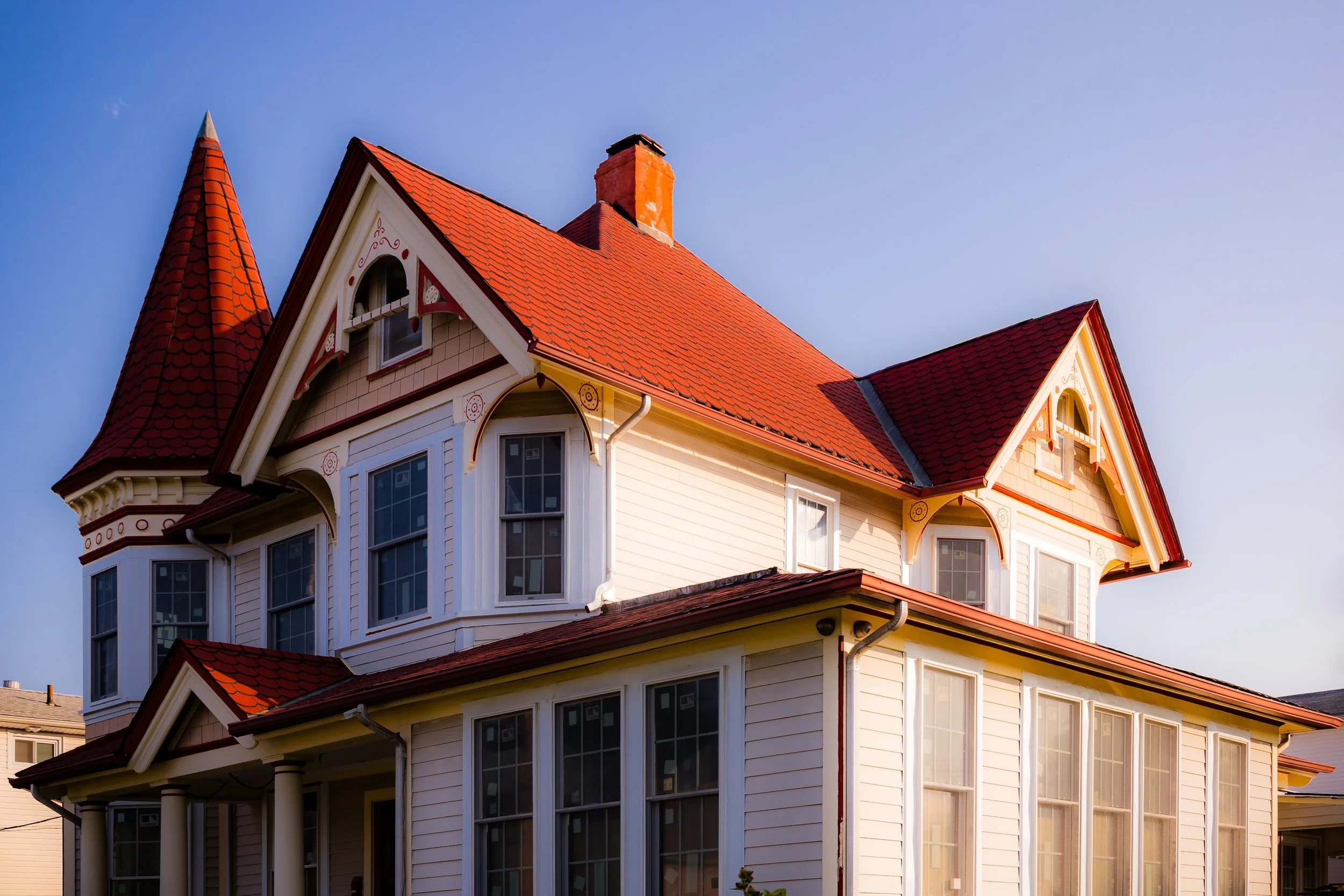Victorian-style house with a red tile roof, large windows, cream siding, and decorative trim, set against a blue sky.
