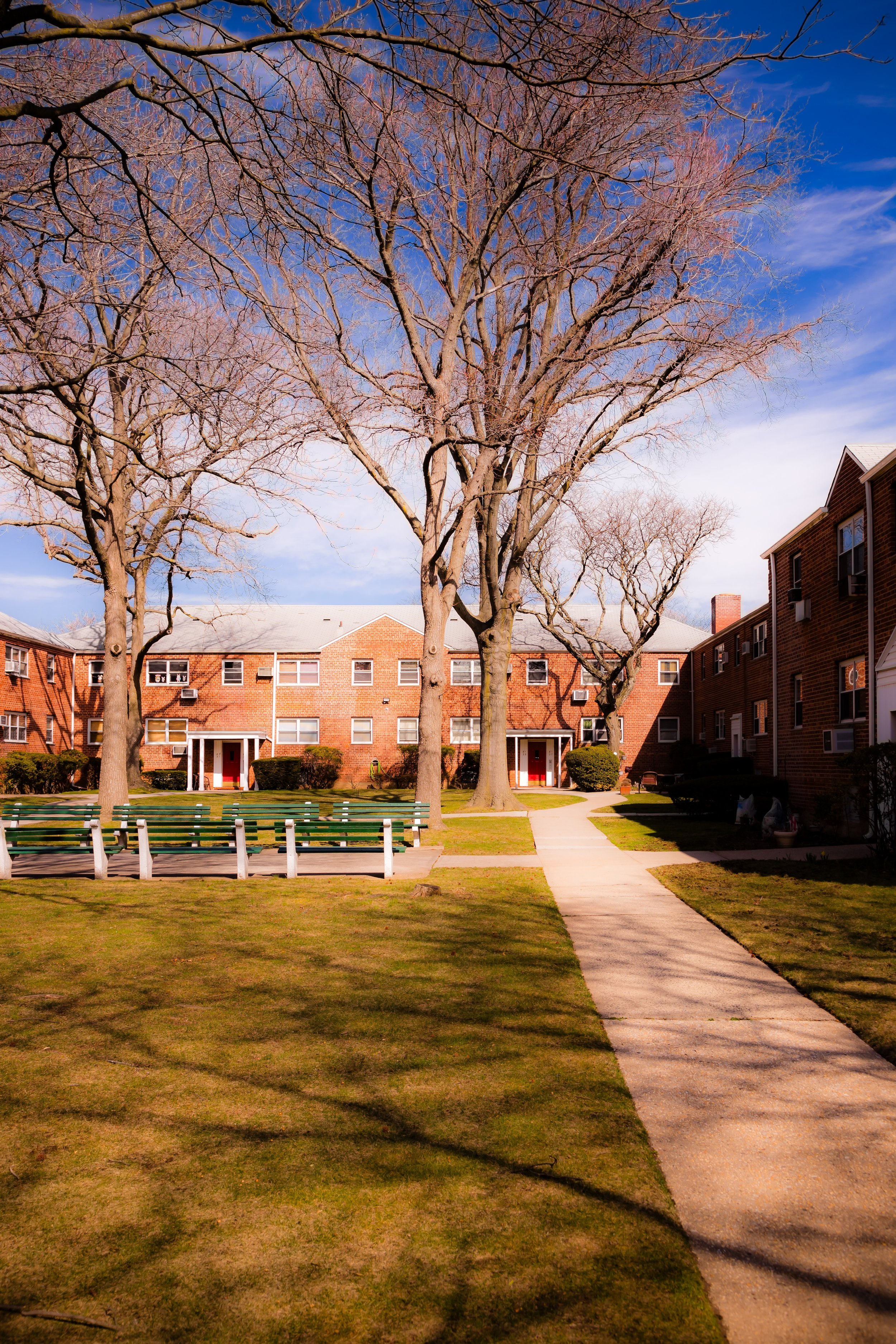A residential courtyard with a concrete pathway, green grass, leafless trees, brick apartment buildings, and empty benches under a blue sky with some clouds.