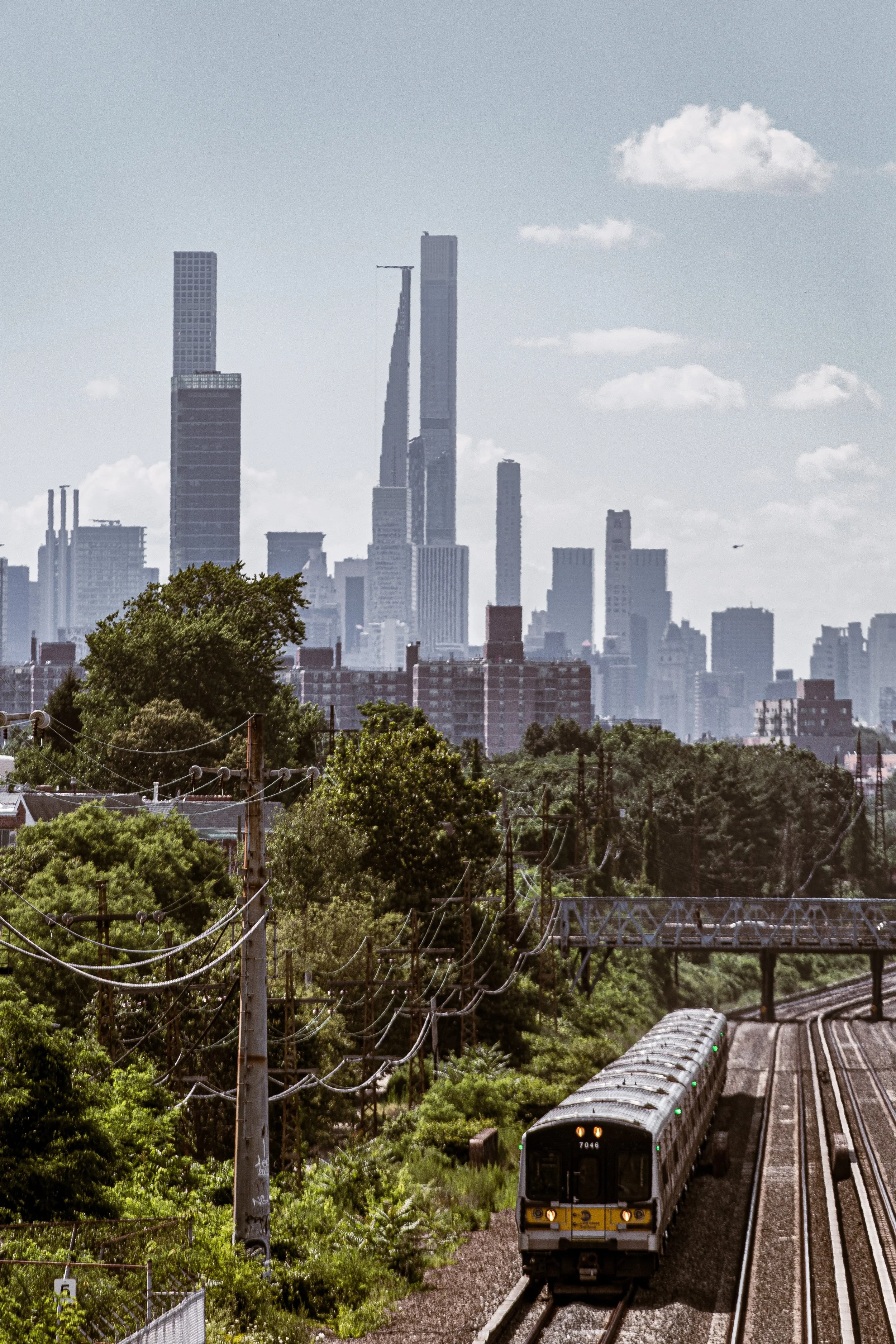 A city skyline with tall skyscrapers in the distance, green trees, and a train on tracks in the foreground.