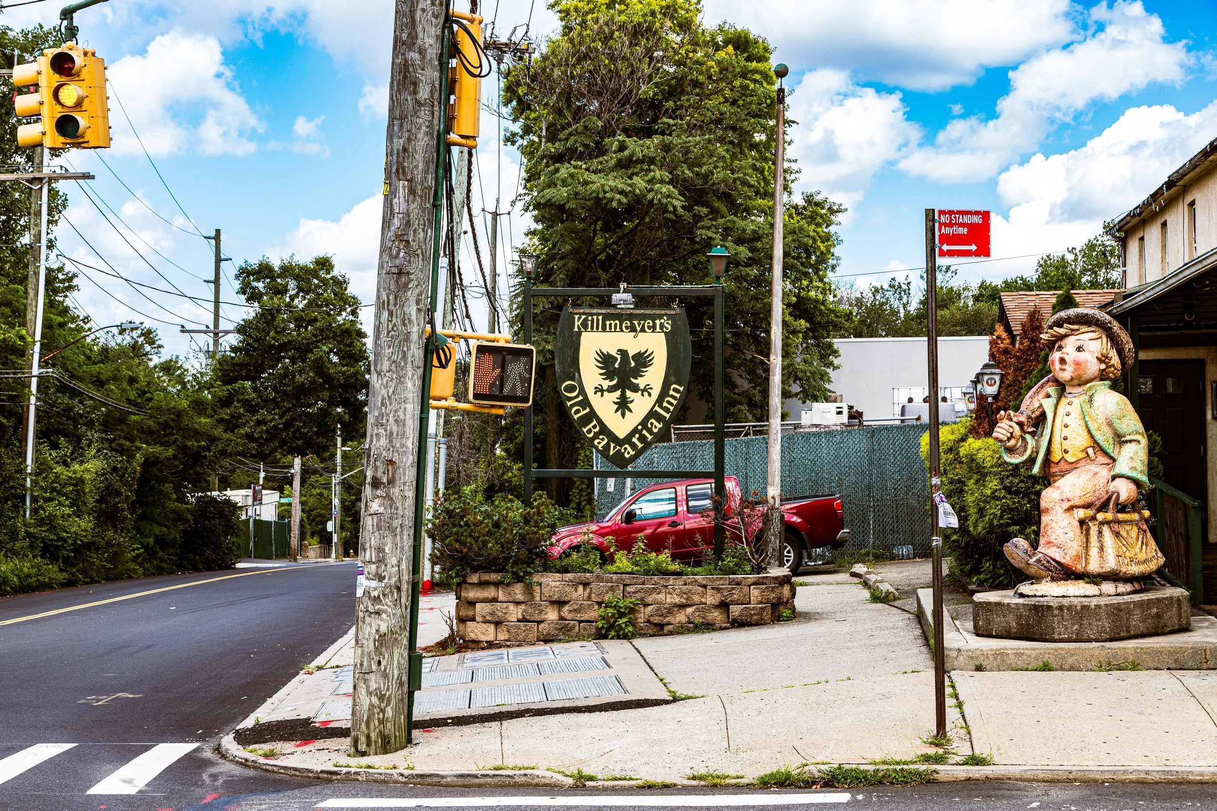 Street view featuring a sign for Killmeyer's Old Bavarian Inn, a decorative sculpture of a boy in traditional clothing, a red pickup truck parked behind a brick planter, a traffic light, and street signs, with trees and a blue sky with clouds in the background.