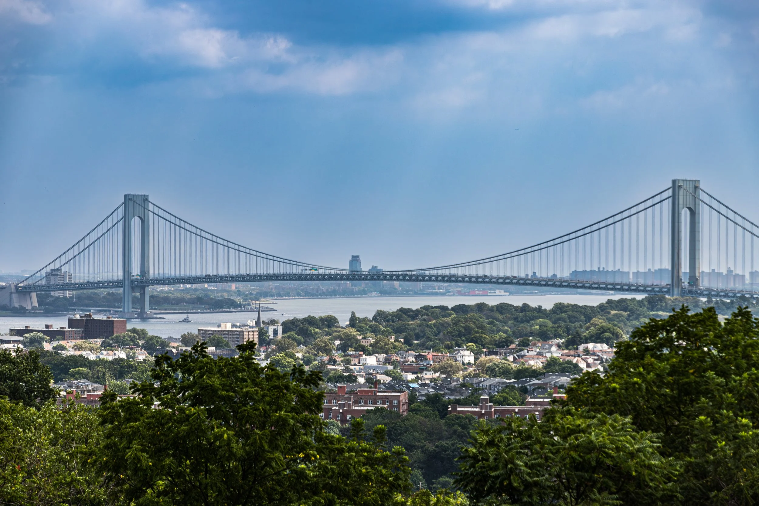 A large suspension bridge over a wide river, with a cityscape in the background and green trees in the foreground.