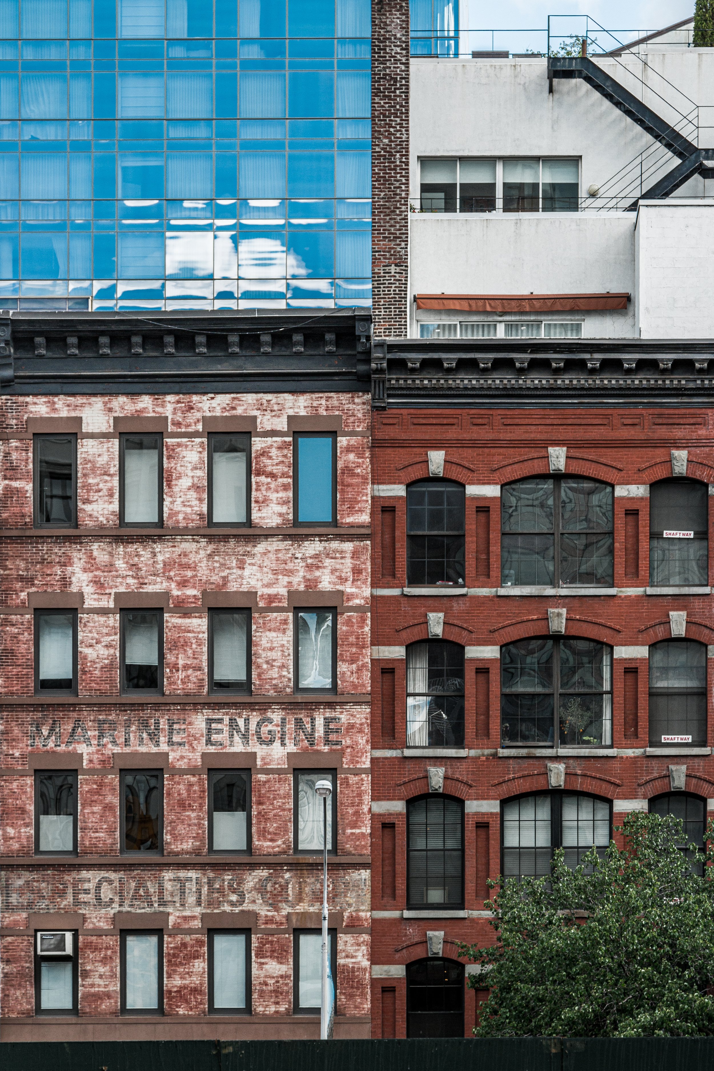 Facade of two adjacent buildings with mixed architectural styles. The left building has a weathered brick exterior with black window frames, and the words 'Marine Engine Mechanics' faintly visible painted on the wall. The right building is a red brick structure with arched windows and decorative stone accents. Part of a modern glass building is visible in the background.