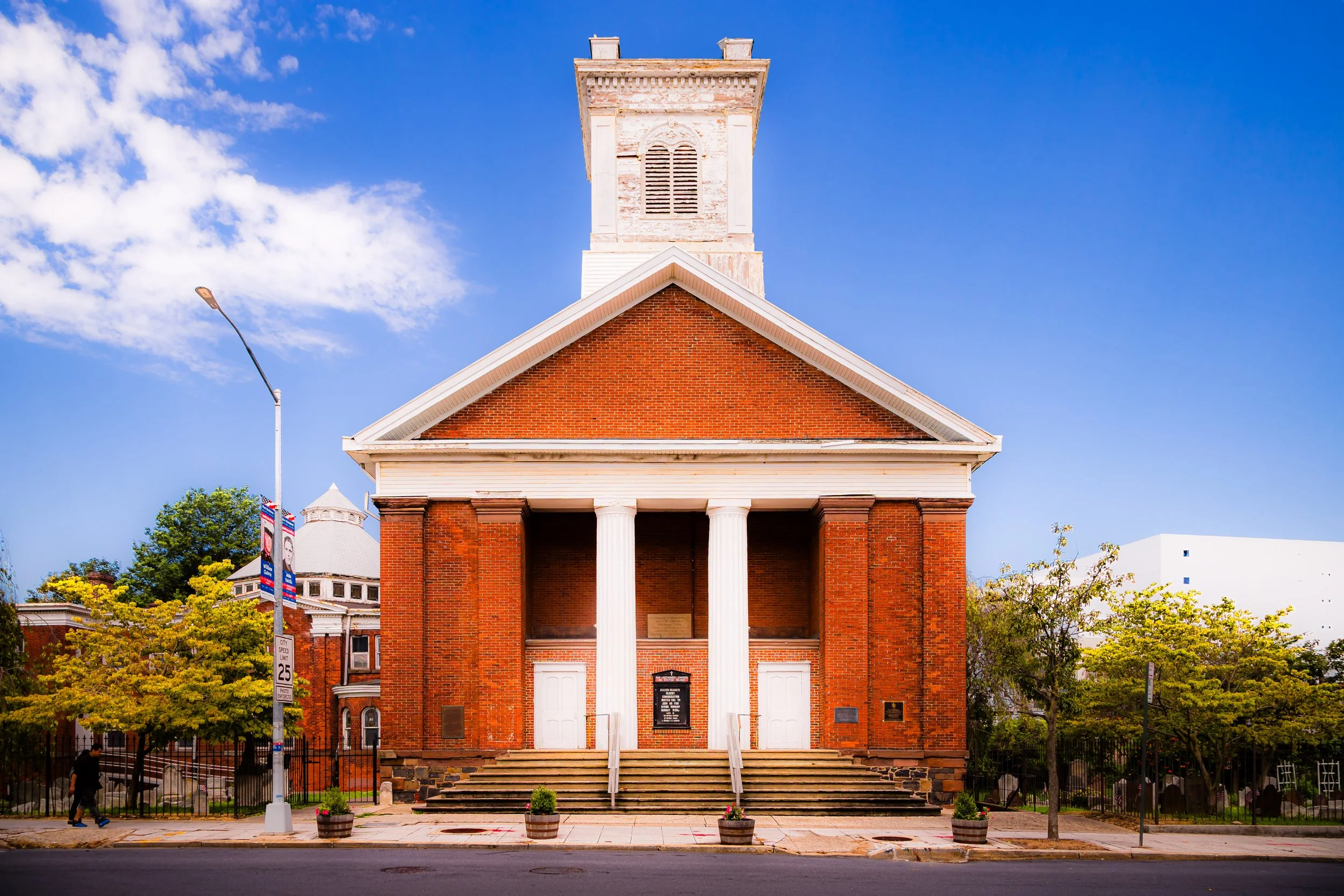 Front view of a red brick church with white columns and a bell tower, set against a blue sky with some clouds. Sidewalk with plants and trees in front.