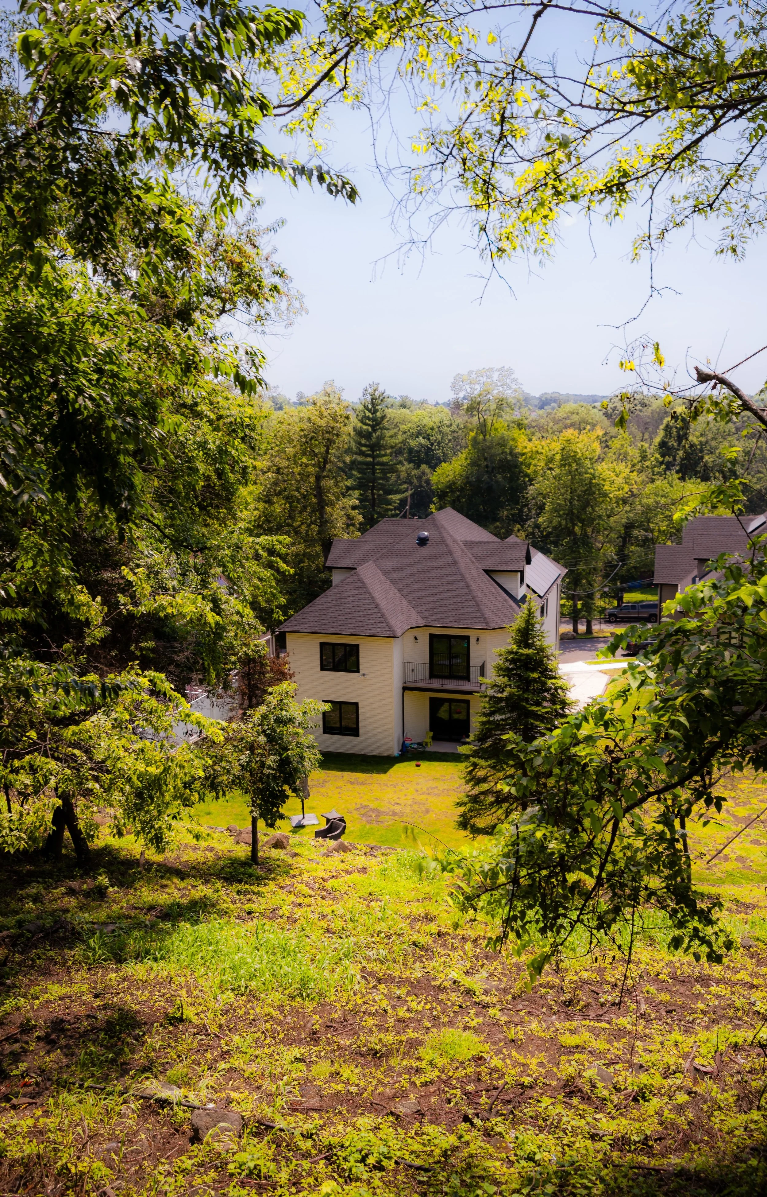 A two-story house with beige exterior walls and a dark brown roof, surrounded by trees and greenery, located in a semi-wooded area with a patch of lawn and patio furniture in the yard.
