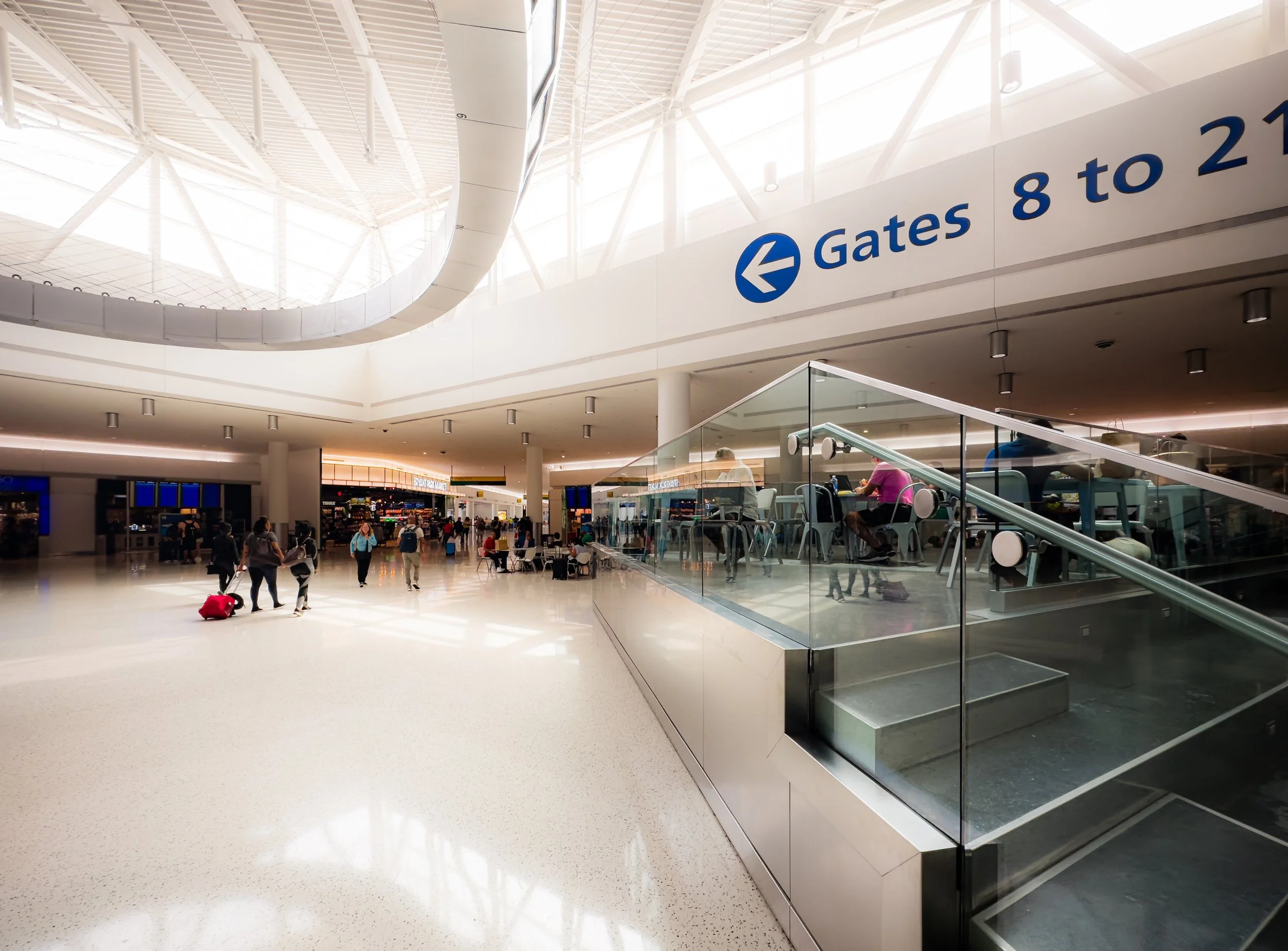 Interior of an airport terminal with people walking and sitting, a sign pointing to gates 8 to 21, and a staircase with glass sides leading downstairs.