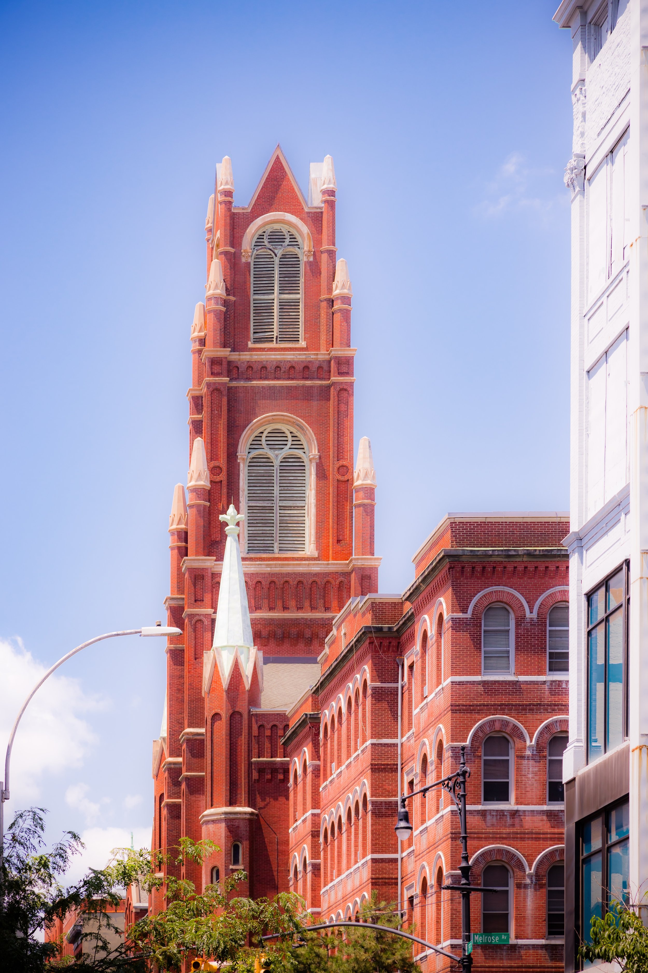 A tall red brick church with gothic architectural features, including arched windows and spires, located on a city street corner with a street sign reading 'Melrose' and a streetlamp in the foreground.