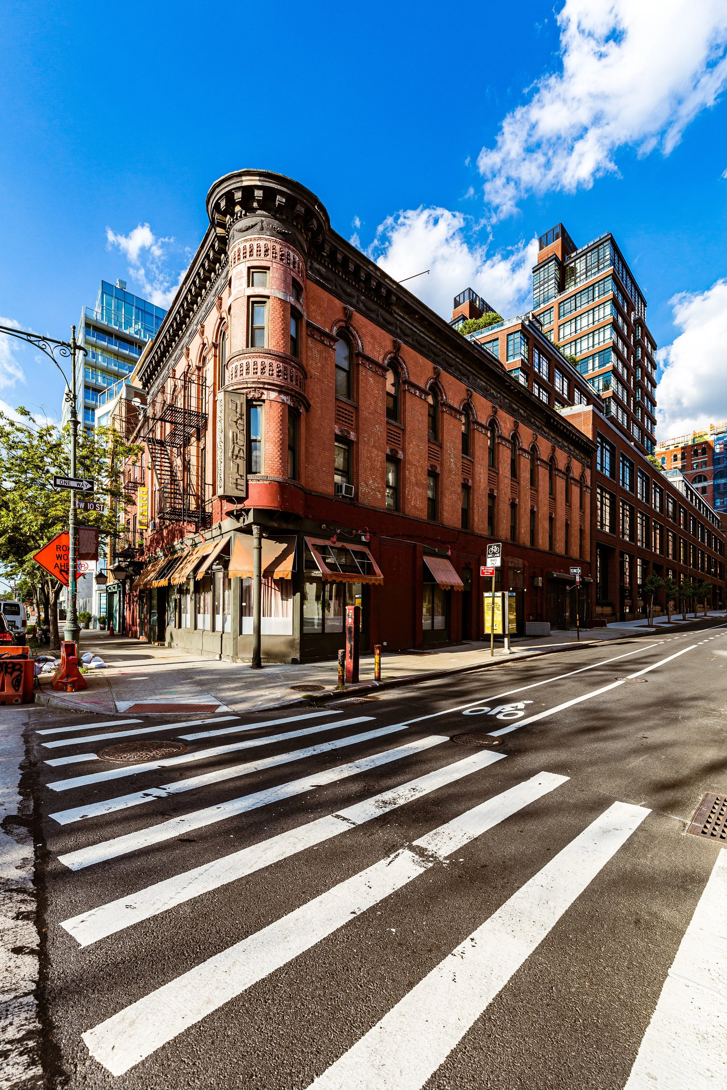 A city street corner with a red brick building on the corner, featuring large windows, awnings, and fire escape stairs, against a bright blue sky with white clouds. Modern high-rise buildings are visible in the background.