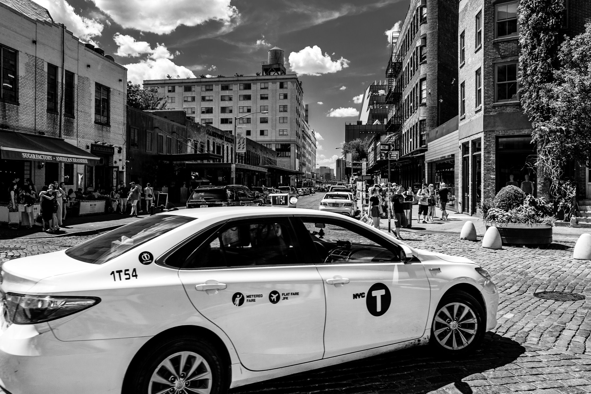 A city street scene in New York City with pedestrians walking on the sidewalk, parked cars, and a white NYC taxi cab in the foreground. Buildings line both sides of the street, and there are clouds in the sky.