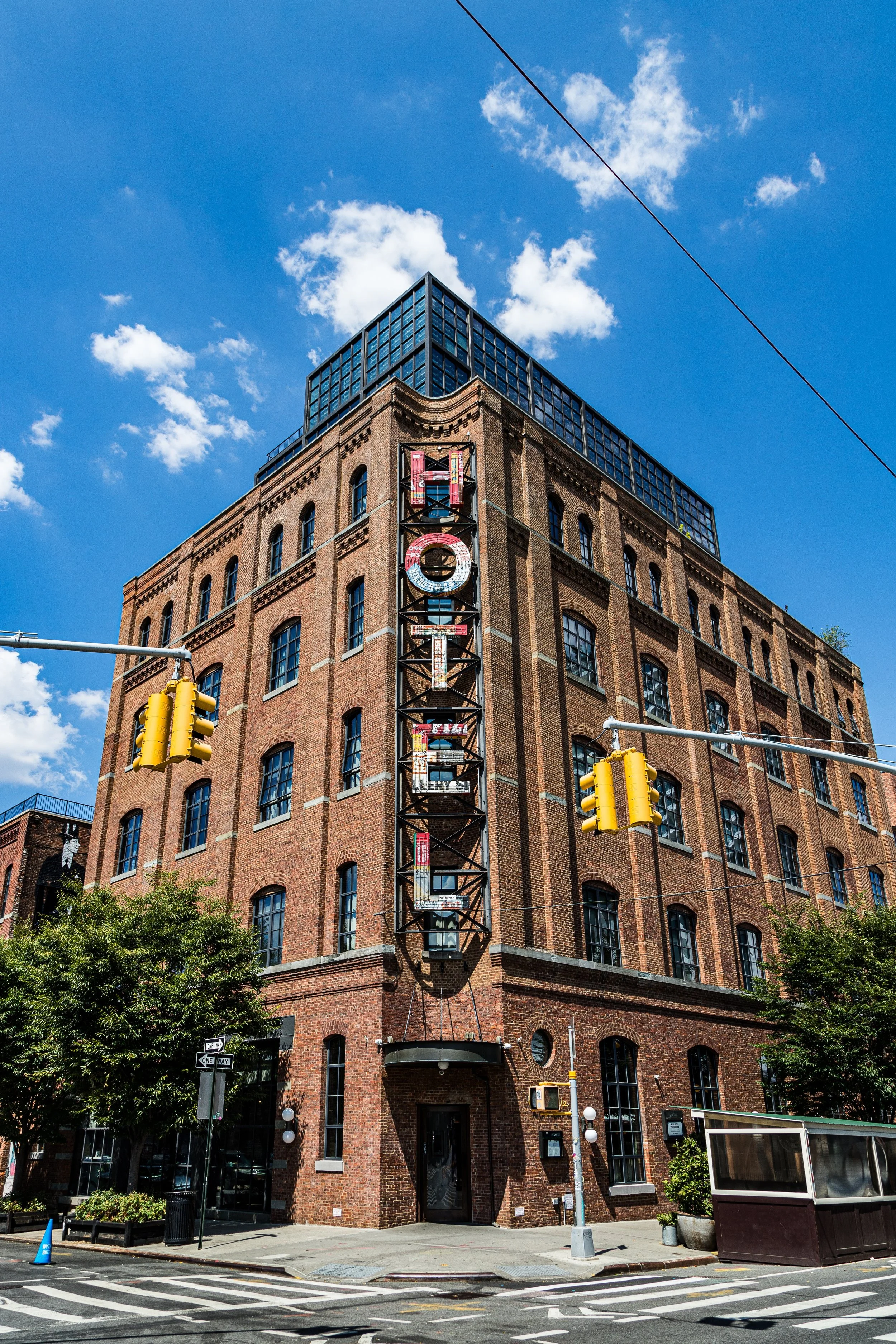 A large brick building with a retro sign reading 'HOTEL' vertically on a black metal structure. The building has multiple arched windows and a modern glass top. The sky is clear with some clouds, and there are traffic lights and trees in front.