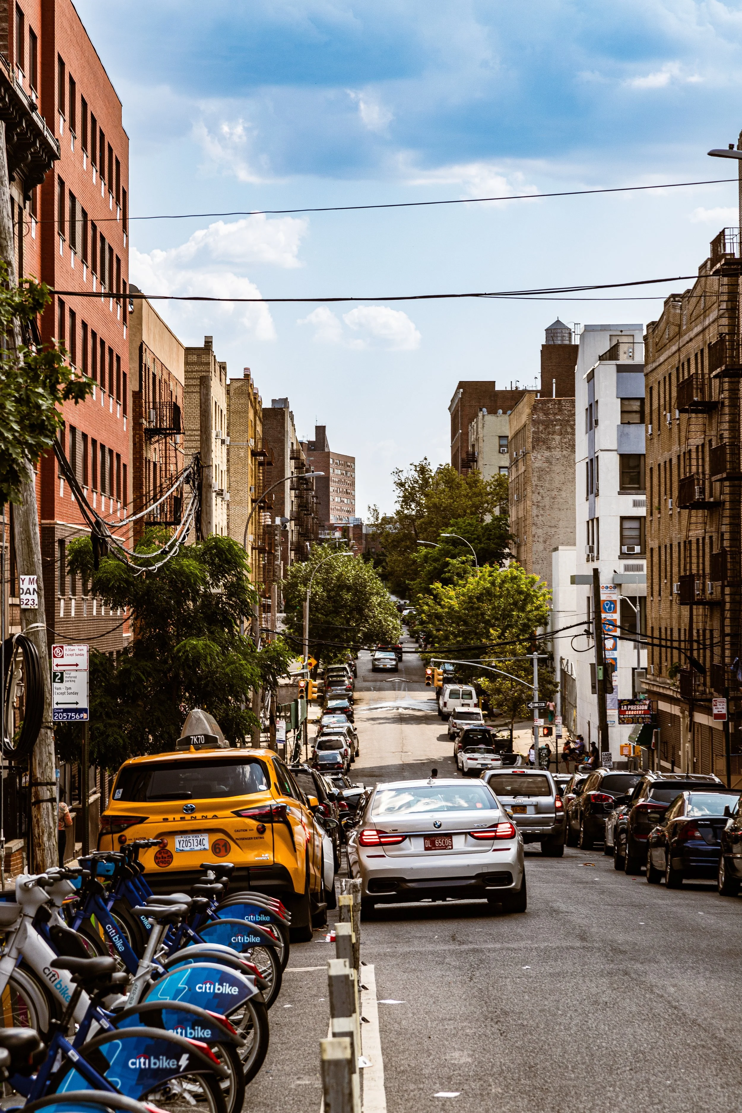 Urban street scene with parked cars and bikes, multi-story buildings, trees, and a blue sky with scattered clouds.