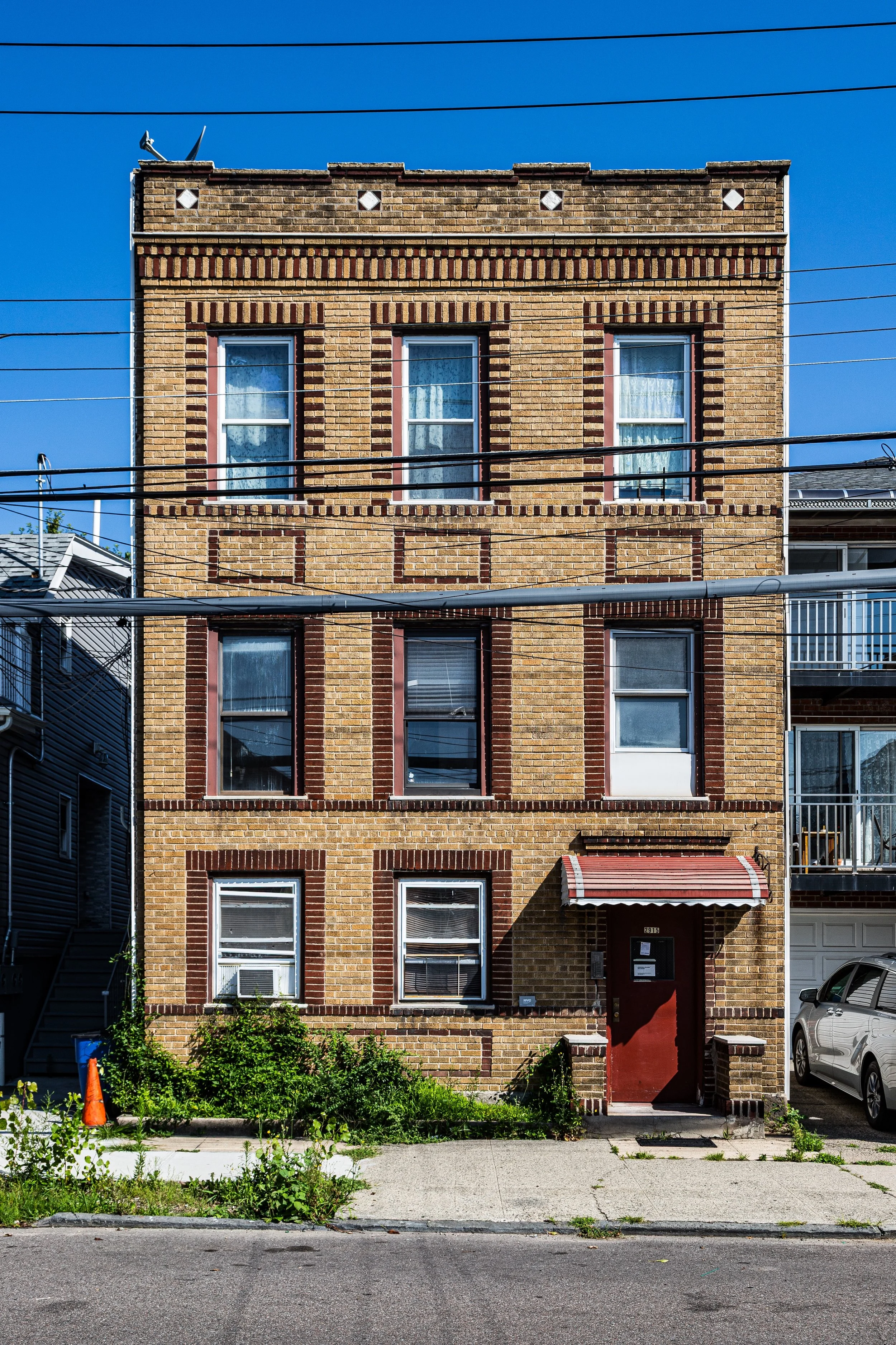 A multi-story brick apartment building with six windows, some with curtains. The building has a red door with a small awning and is surrounded by some plants and parked cars, with a clear blue sky overhead.