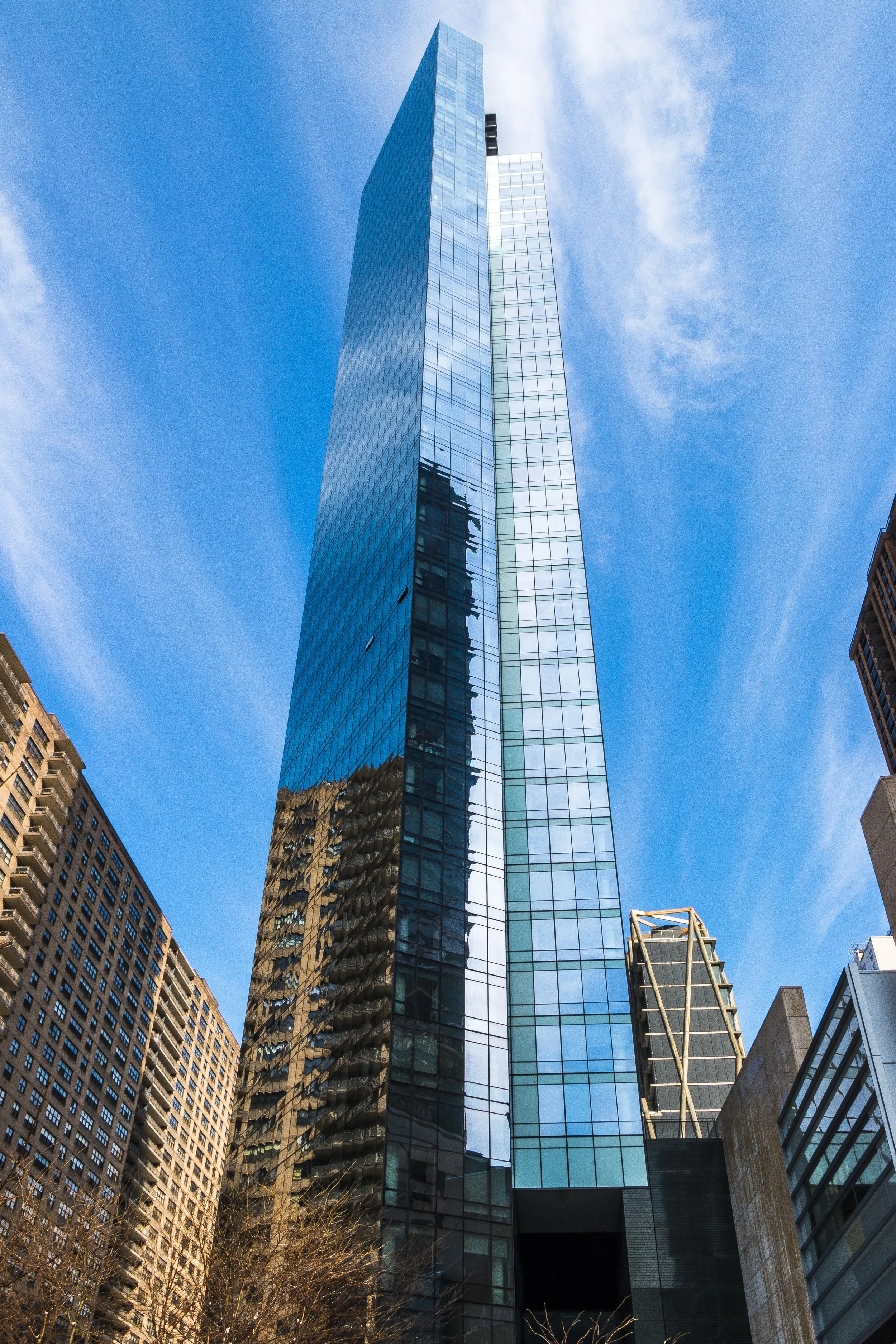 Tall blue glass skyscraper towering over surrounding buildings in an urban cityscape under a partly cloudy sky.