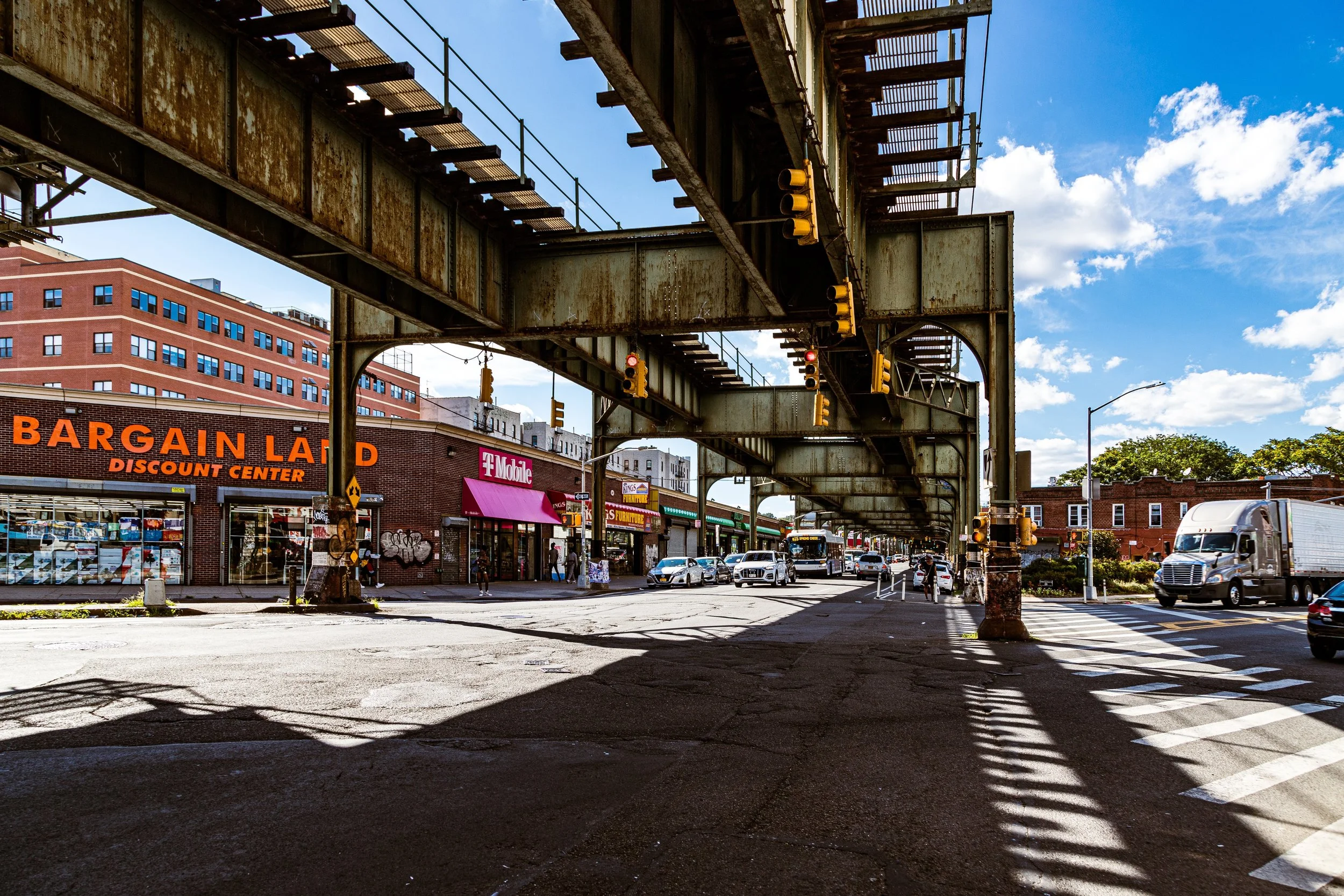 City street scene with an elevated train track overhead, traffic signals, cars, a delivery truck, and storefronts including Bargain Land Discount Center and T-Mobile, on a sunny day with blue sky and some clouds.