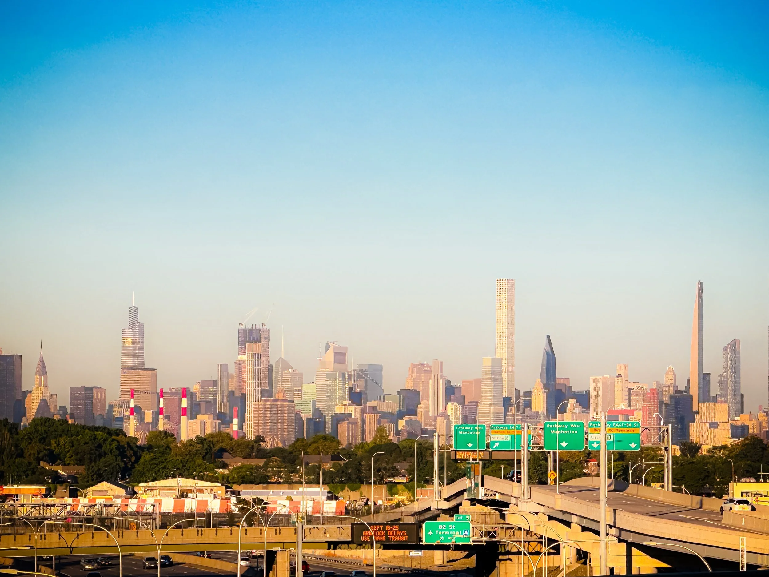City skyline of New York City with tall skyscrapers, visible highway with green overhead signs, and some vehicles in the foreground during daytime.