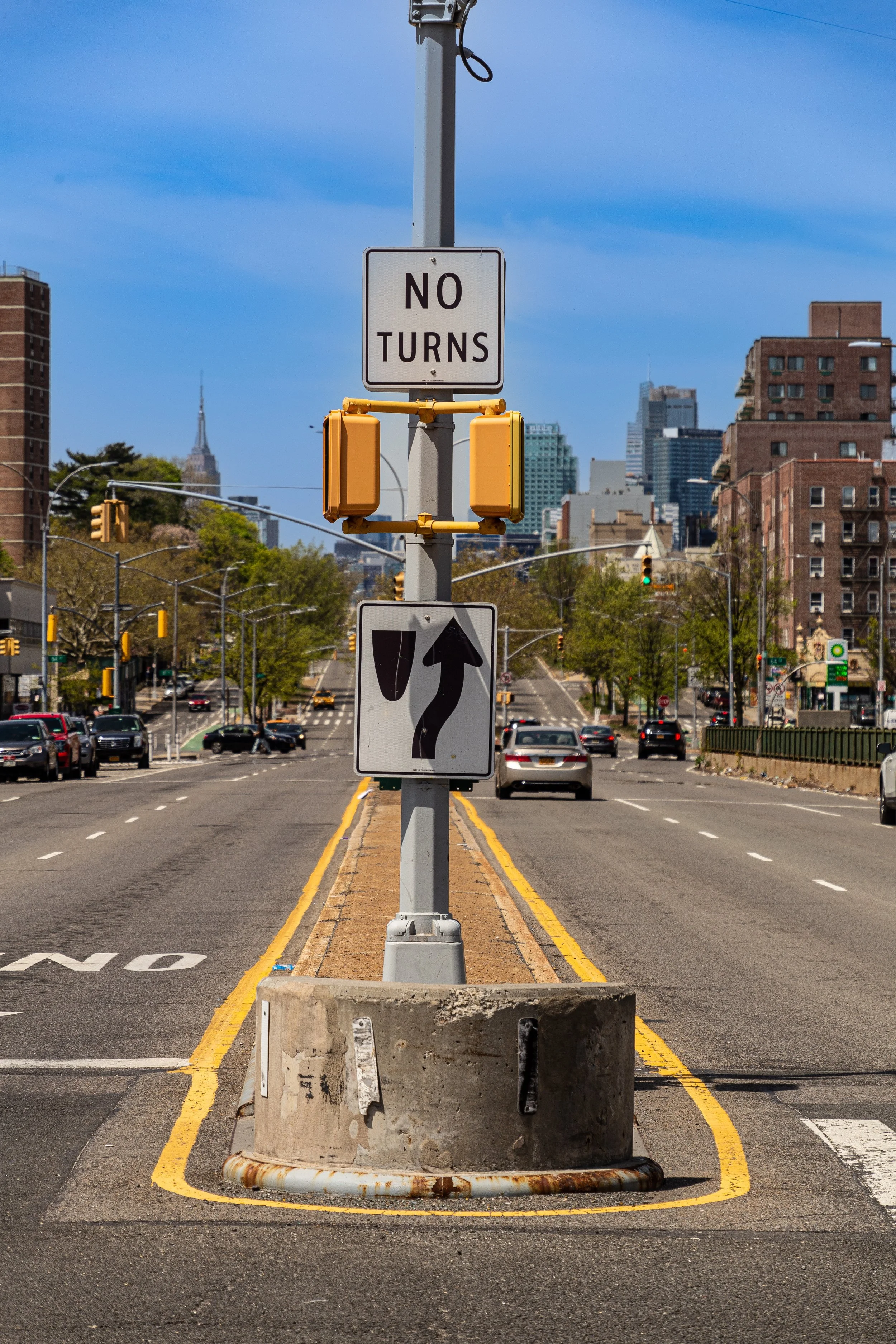 City street with traffic, buildings, and a pole with signs indicating no turns and a one-way arrow pointing straight.