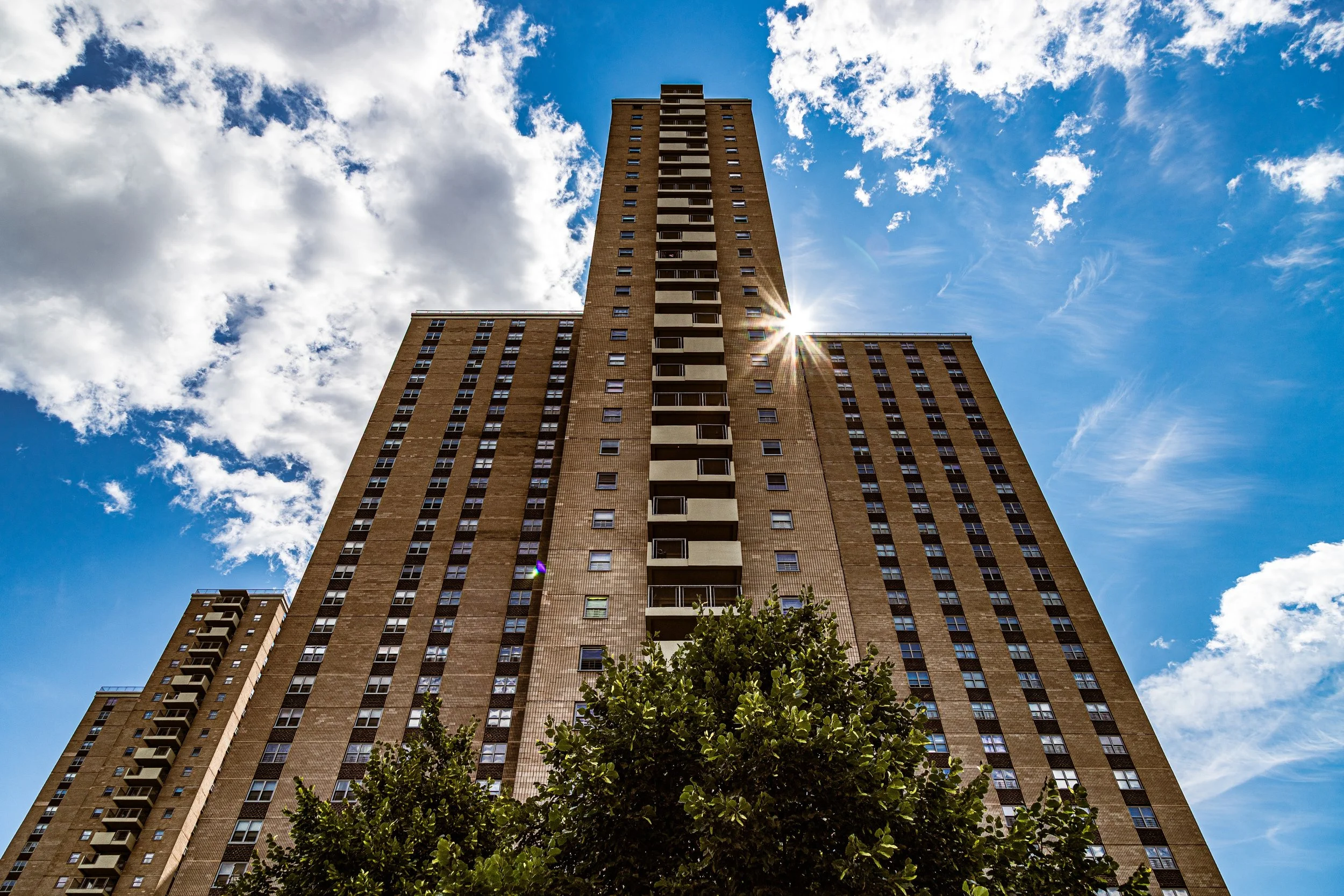 Low-angle view of a tall apartment building against a bright blue sky with scattered clouds and sunlight reflecting off the building.
