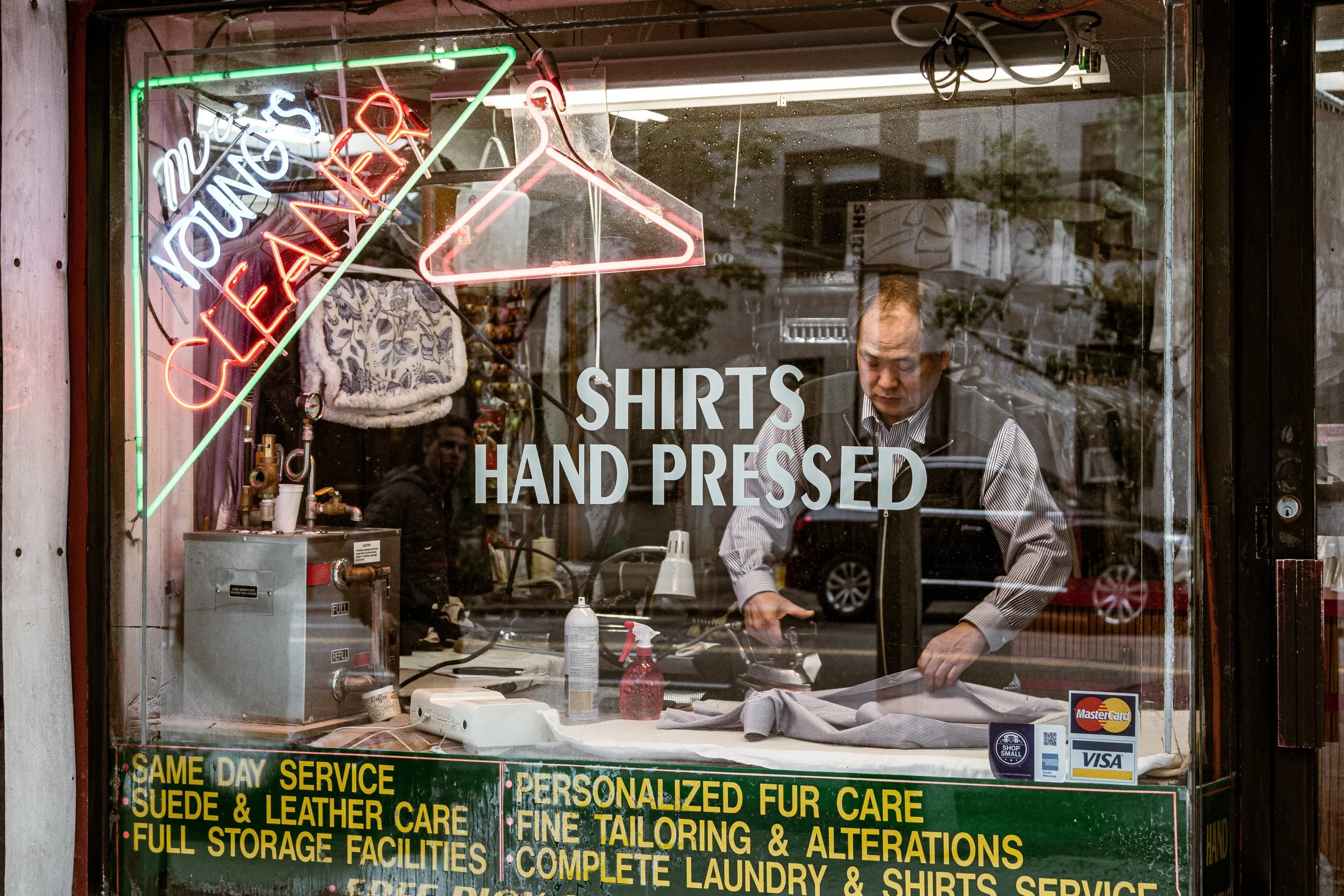 A man working inside a shirt printing shop, seen through a glass window with signs and neon lights. The window displays the phrase 'SHIRTS HAND PRESSED' and advertising for same-day service, suede and leather care, personalized fur care, full storage facilities, and laundry and shirt services.