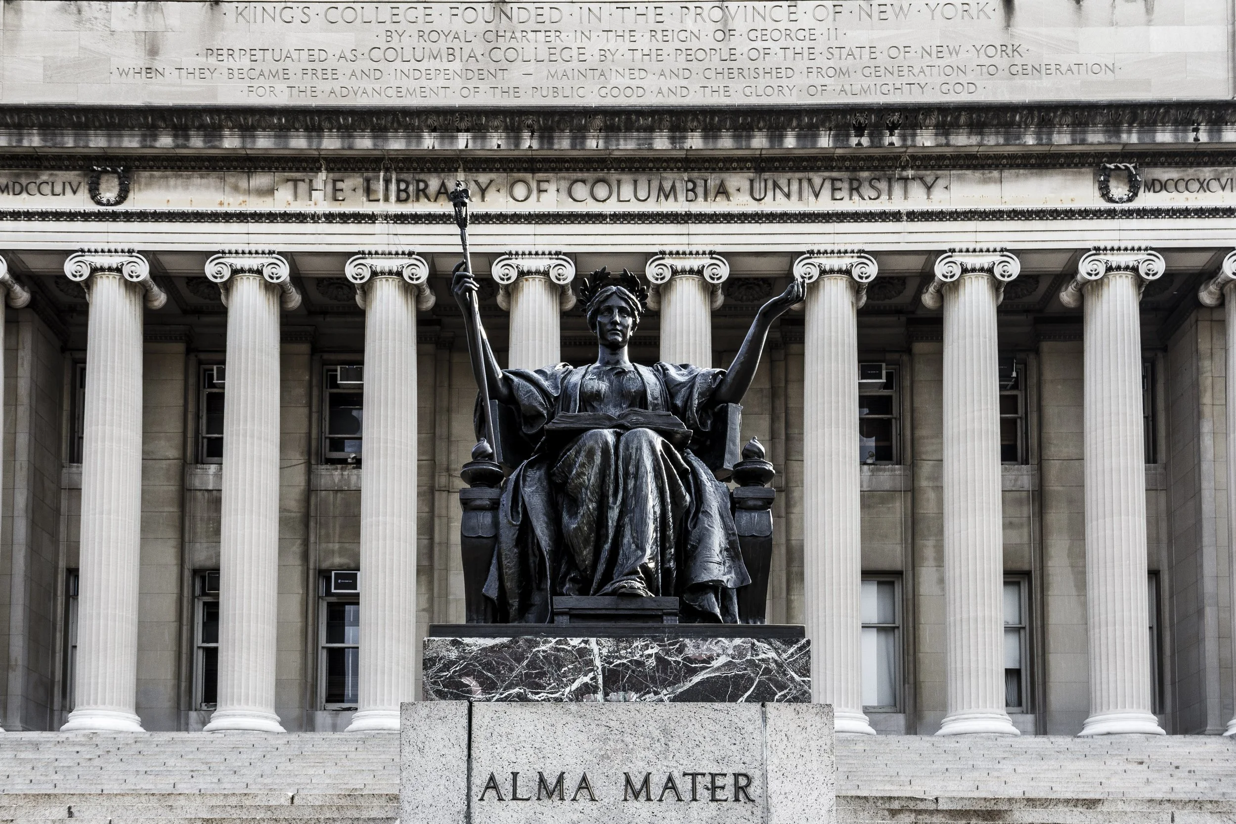 Bronze statue of a seated woman holding a scepter and book, with one hand raised, in front of the Columbia University library building with tall columns and an inscription reading 'THE LIBRARY OF COLUMBIA UNIVERSITY'.