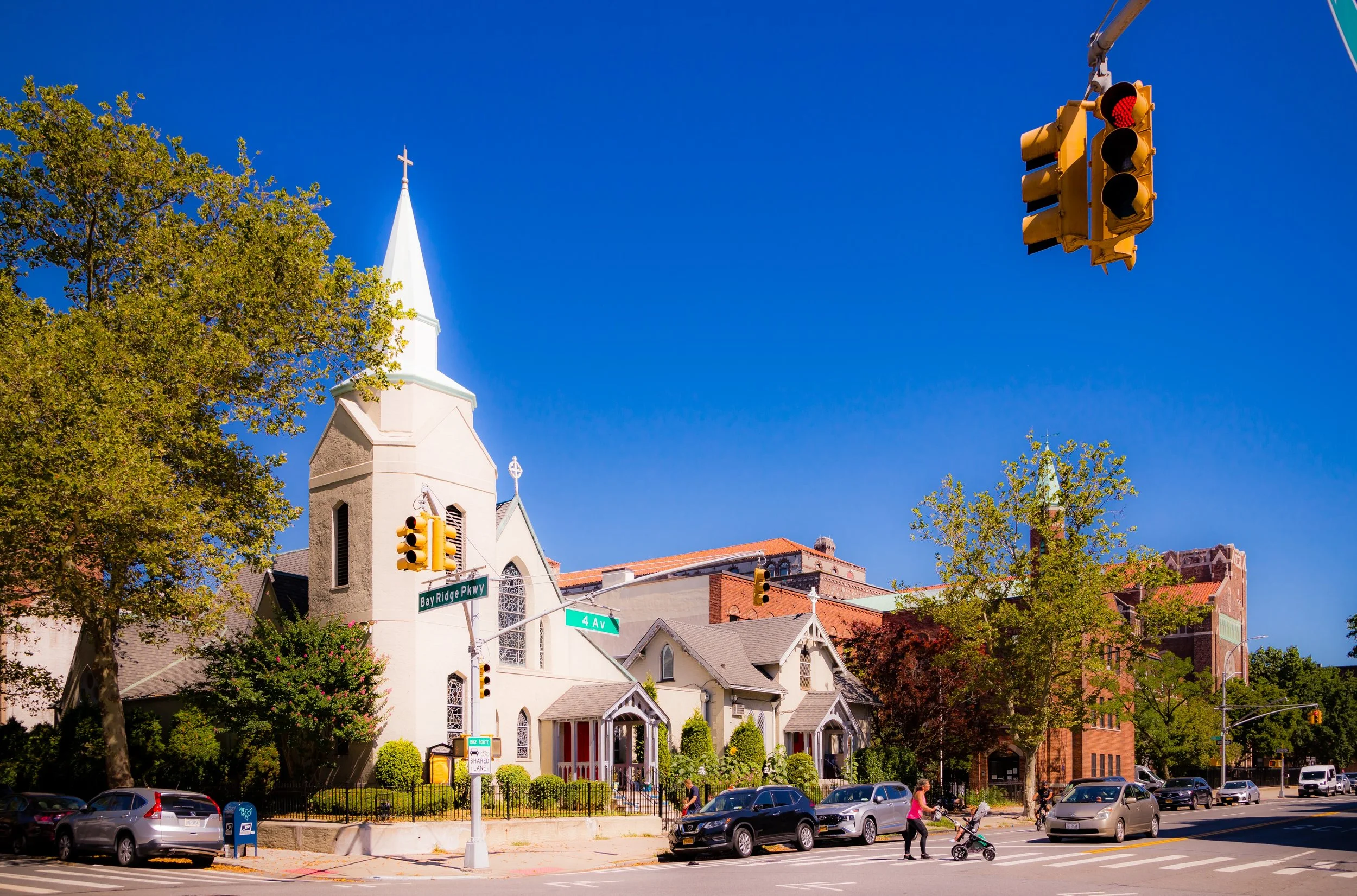 A church at an intersection with traffic lights and cars parked along the road on a sunny day, with trees and a blue sky in the background.