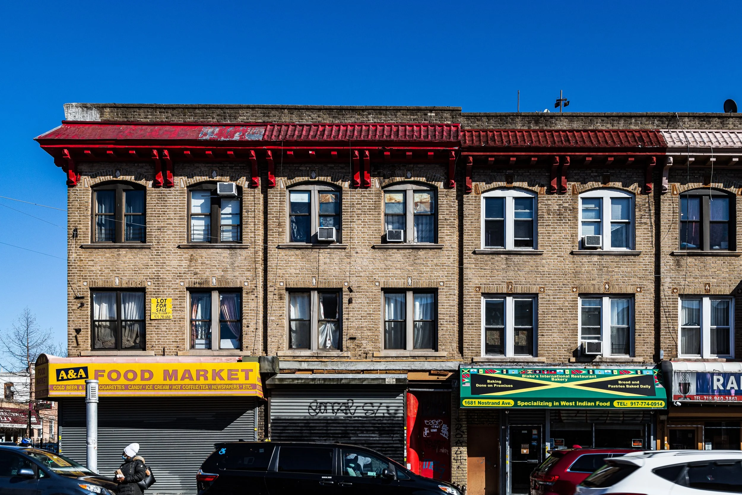 Three-story brick building with storefronts on the ground floor, including A&A Food Market and a bakery. Several cars parked in front, a person walking, blue sky above, and windows with air conditioning units on the upper floors.