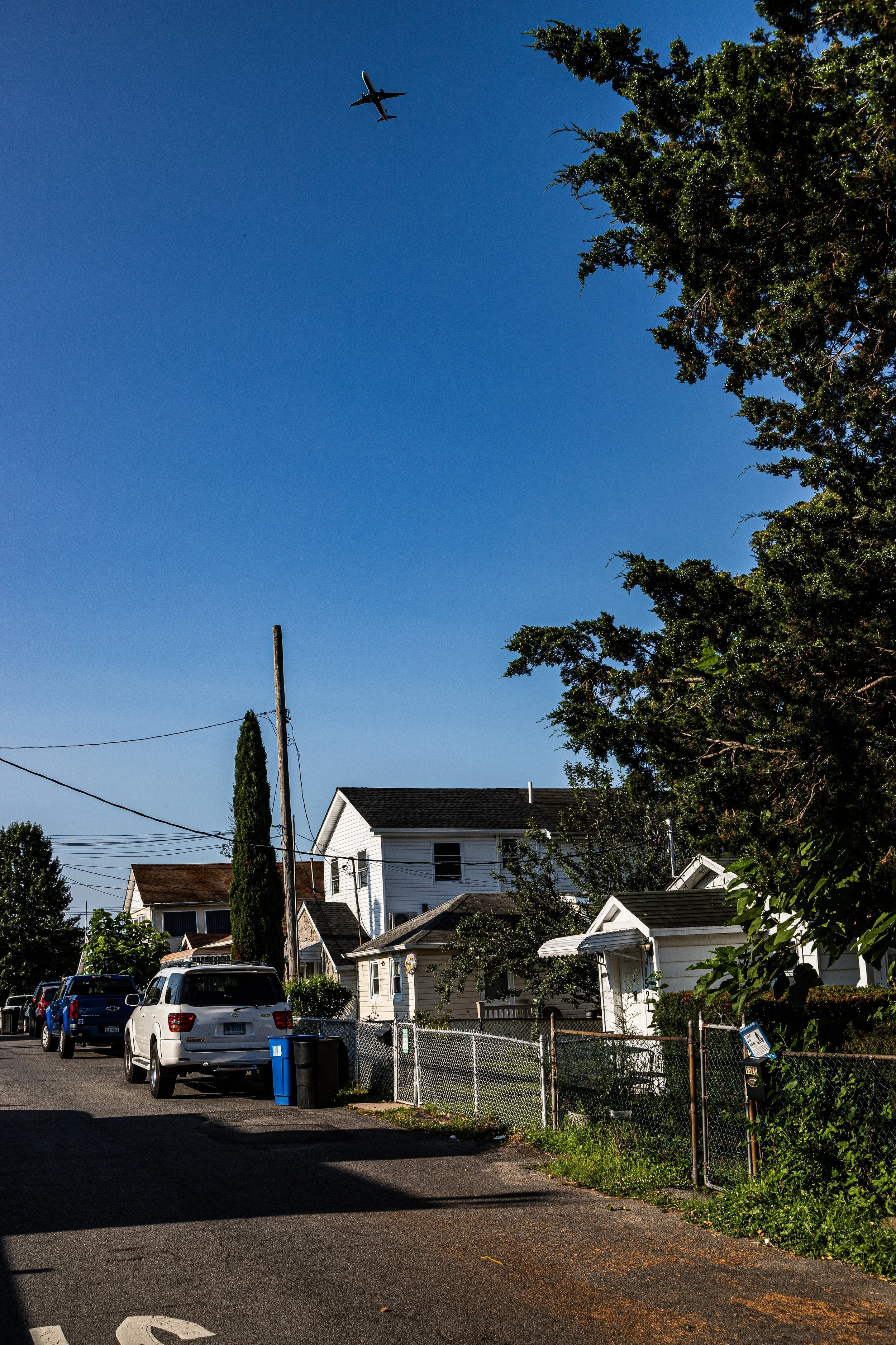 Residential neighborhood with white houses, parked cars, and a utility pole, under a clear blue sky with an airplane flying overhead.