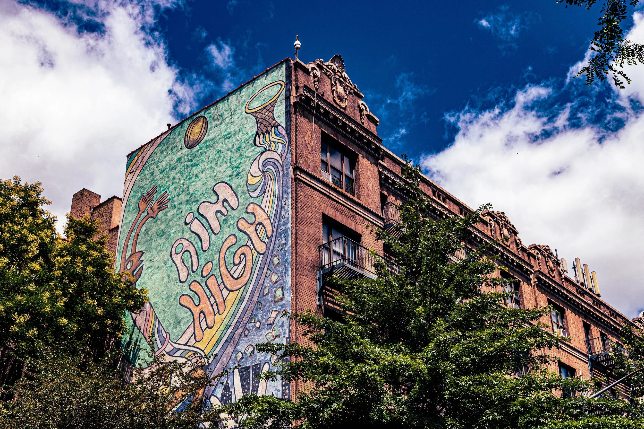 A mural on a brick building features hands reaching up and swirling designs with the words "Paint Arcade". Trees are in the foreground under a partly cloudy blue sky.