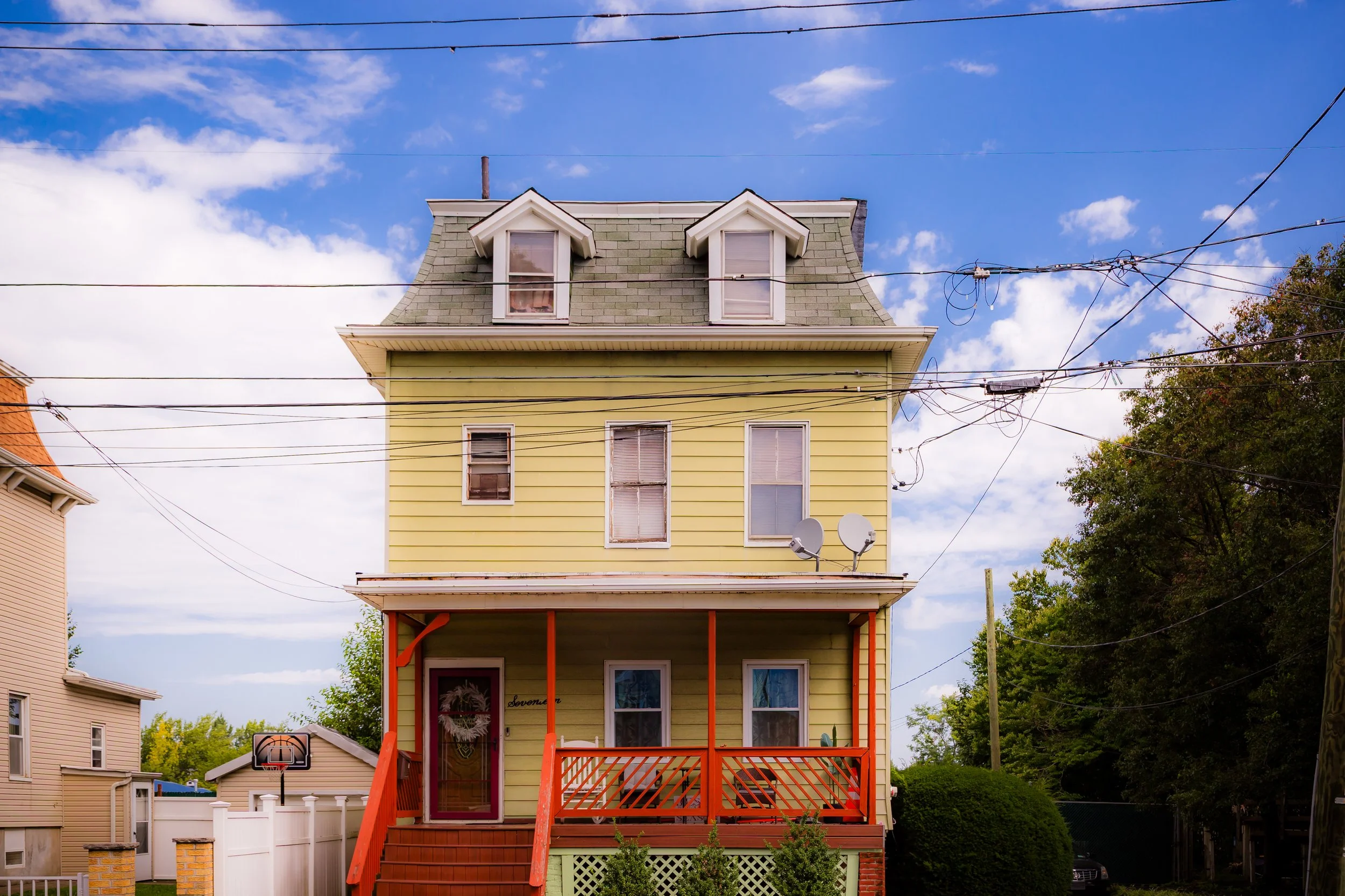 A three-story yellow house with gray dormer windows and a covered front porch with orange railings and steps, surrounded by greenery and other houses, under a partly cloudy sky