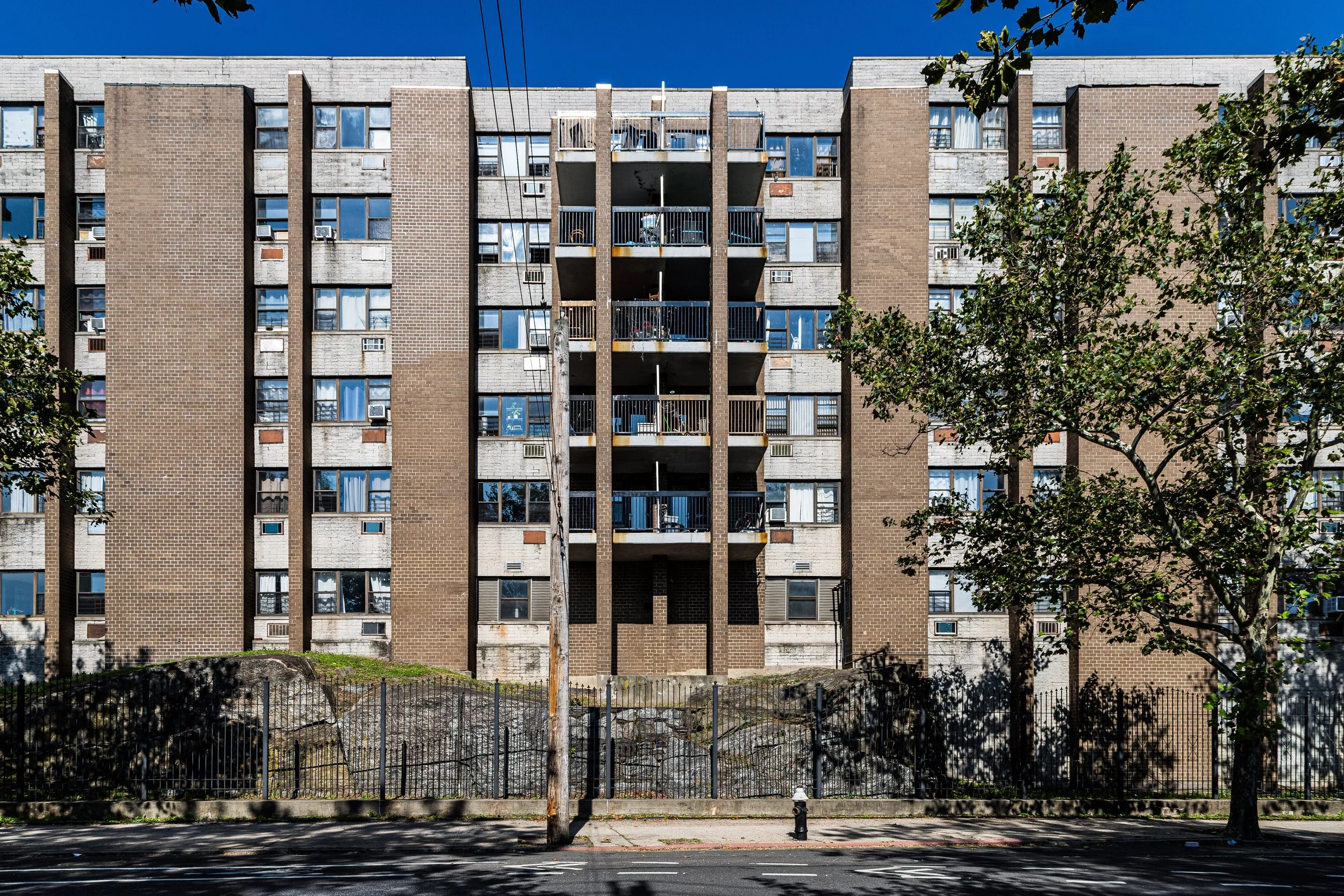 Front view of a multi-story residential apartment building with brick and concrete facade, multiple windows, and balconies, with trees and a sidewalk in the foreground.