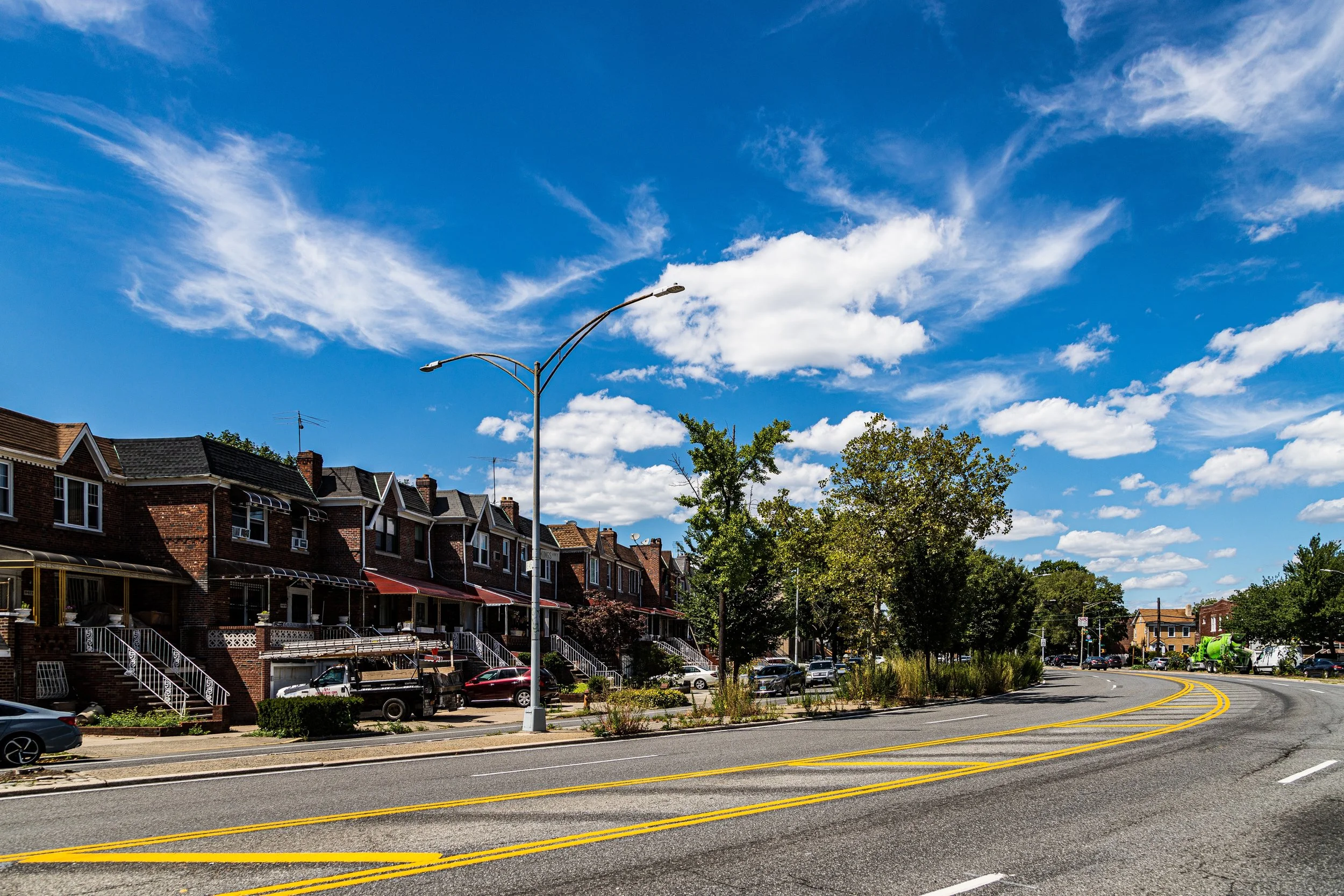 Residential neighborhood street with brick houses, parked cars, trees, and a bright blue sky with scattered clouds.