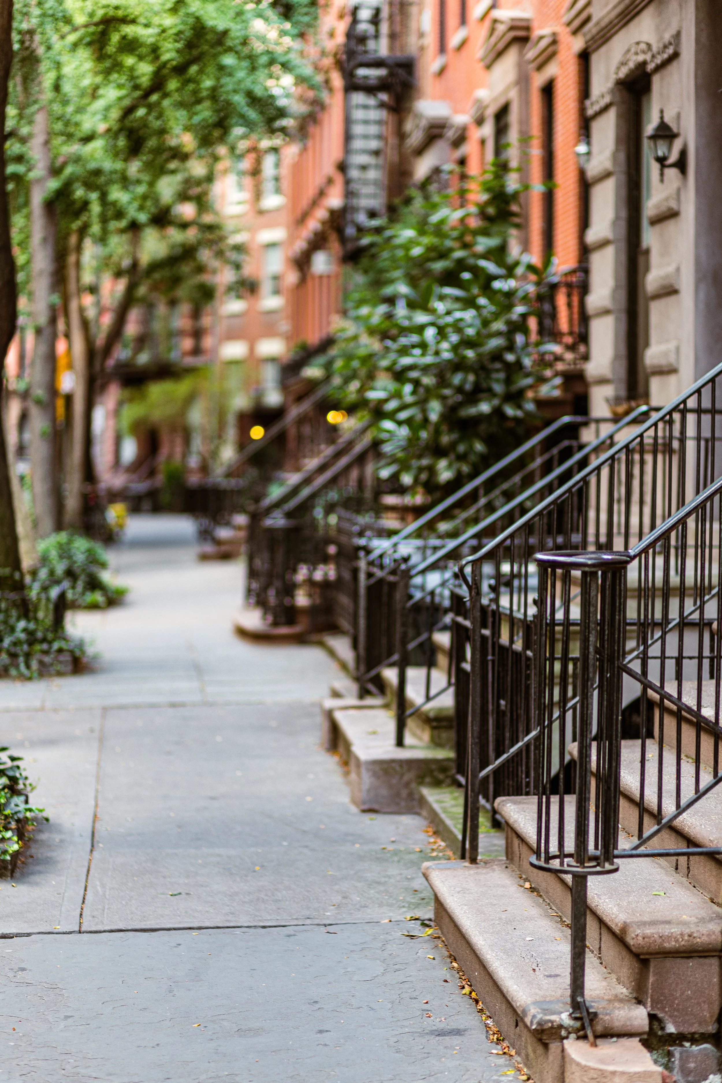 A sidewalk in an urban neighborhood with brownstone buildings, some with stairs leading up to their entrances, black metal railings, and trees lining the street.