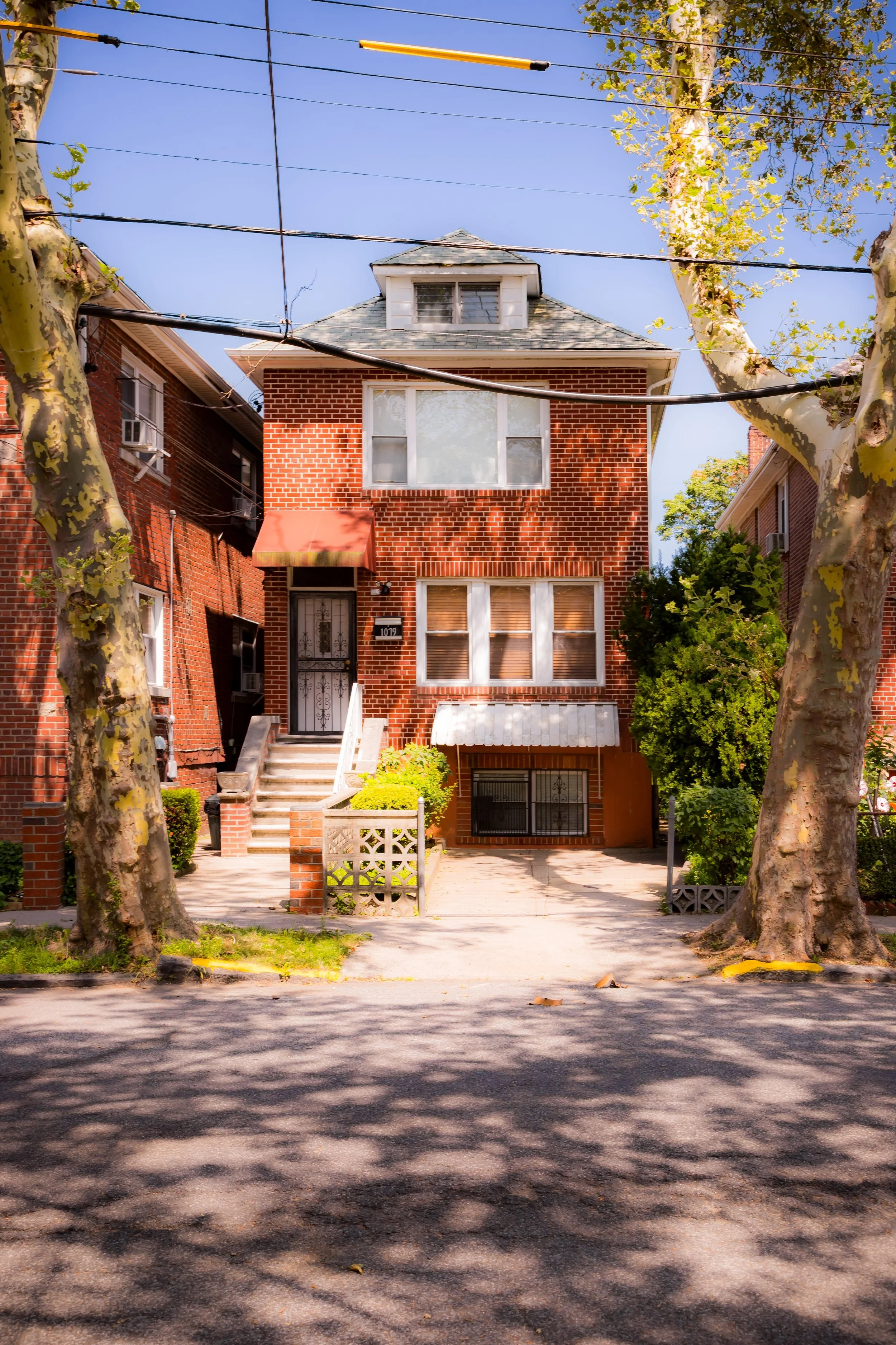 A three-story brick house with stairs leading to the front door, surrounded by large trees on a sunny day.