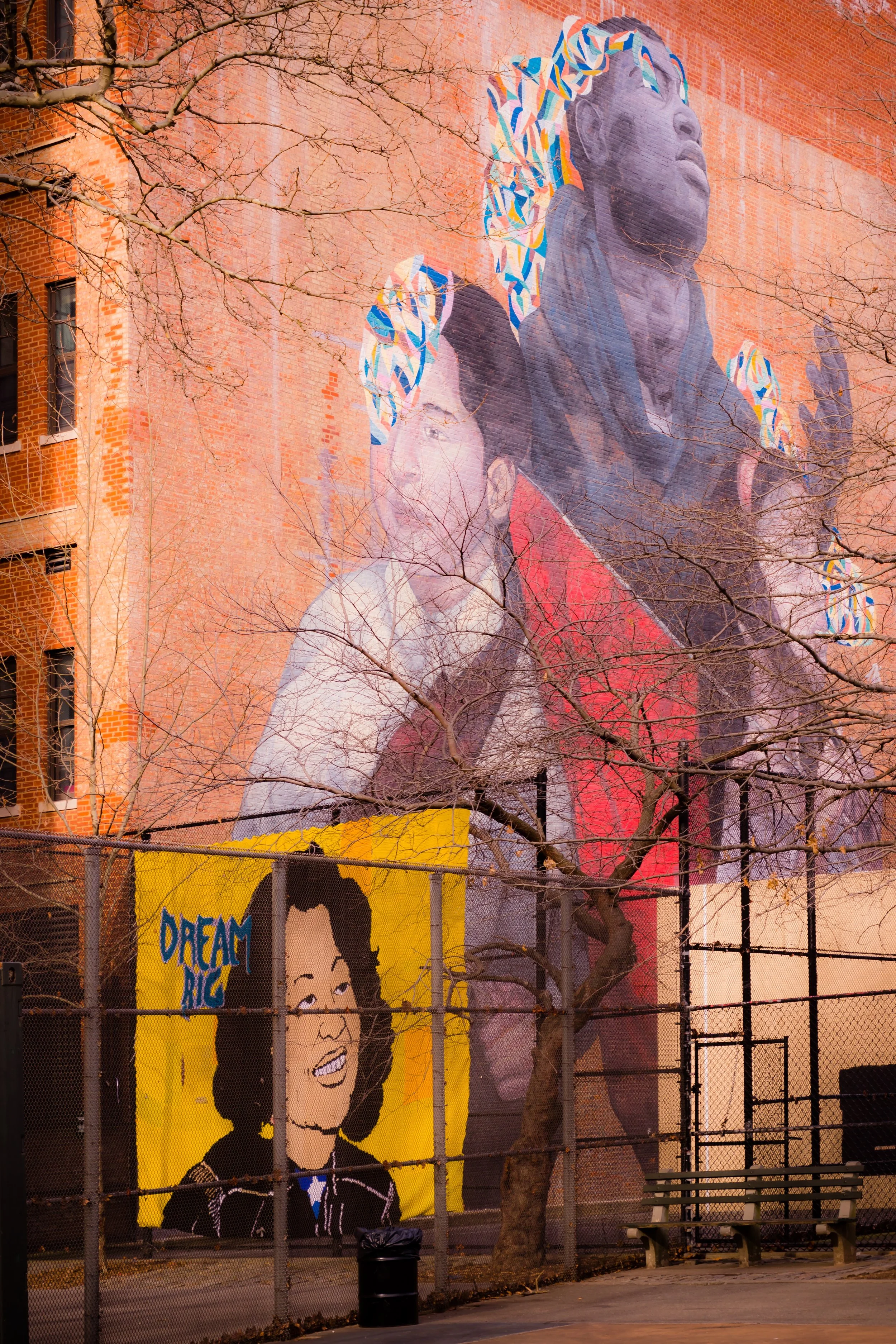 A large mural on a brick wall featuring a woman and a gorilla, with colorful geometric patterns on the gorilla's head. In the foreground, there is a smaller painting of a smiling woman with the words "DREAM BIG" on a yellow background, placed next to a park bench and a tree with bare branches.