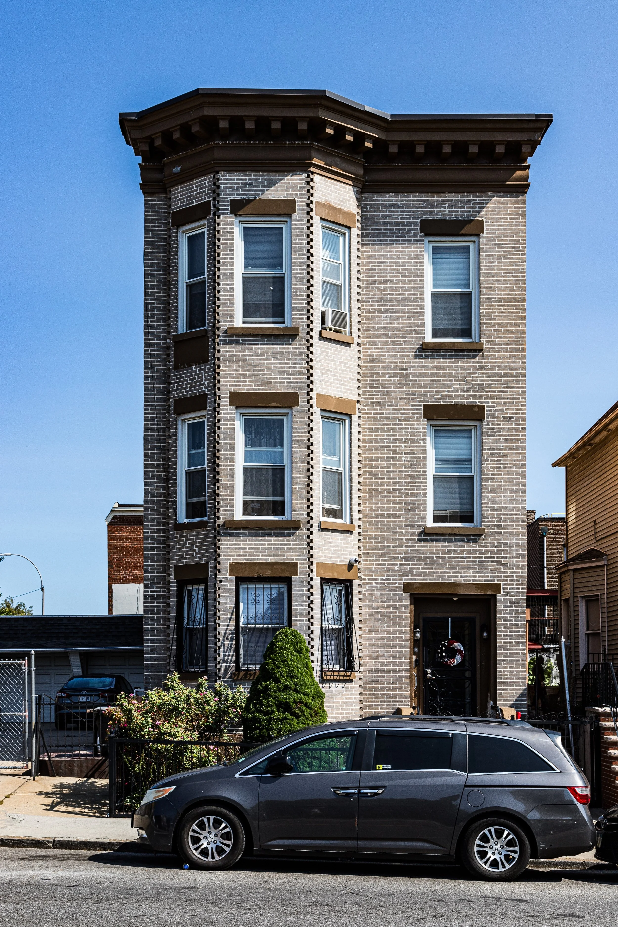 A three-story brick residential building with bay windows and a black front door, decorated with a holiday wreath. A black minivan is parked in front on the street, and the scene includes some greenery and flowers near the entrance.