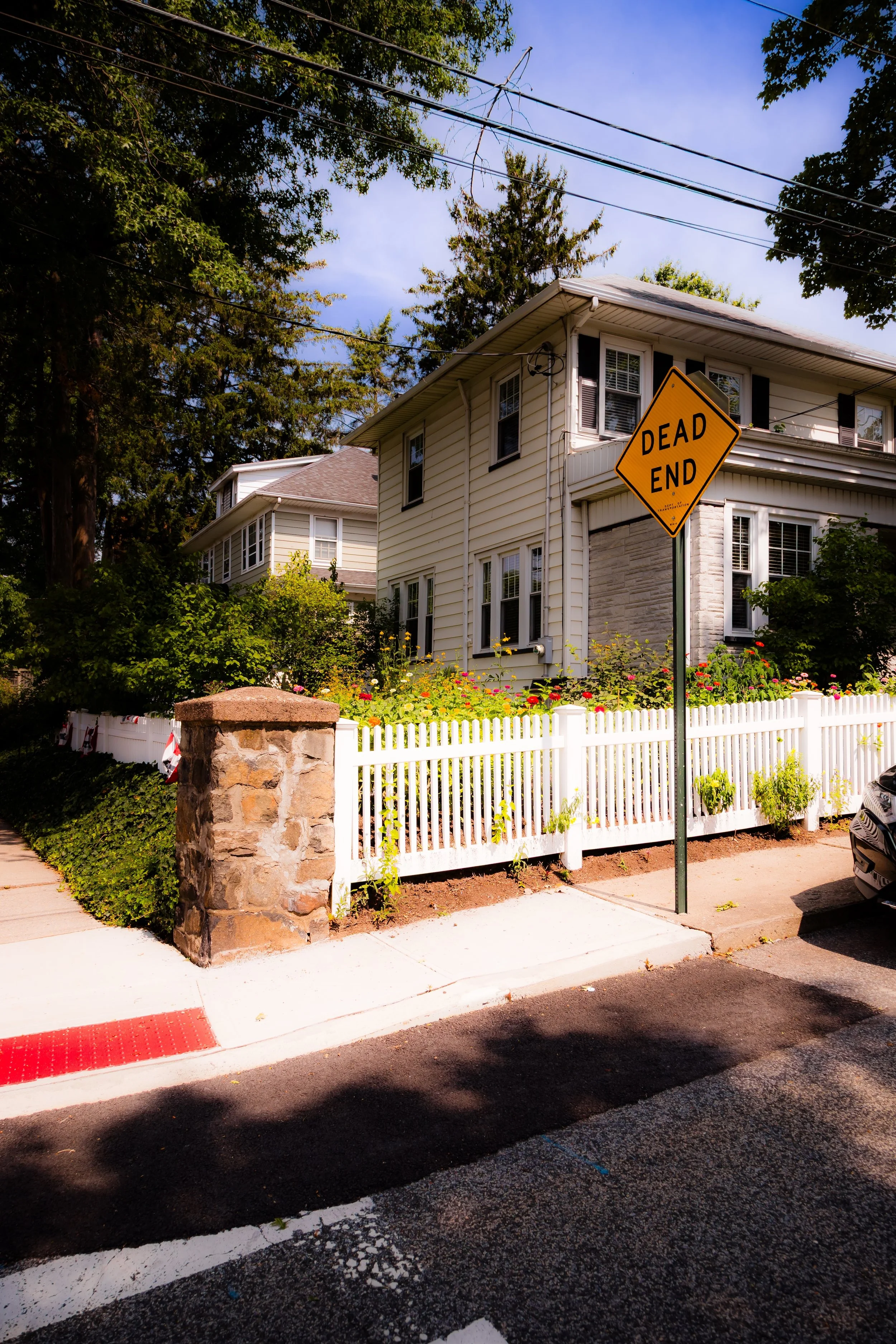 Residential neighborhood street with a white picket fence, a stone pillar, and a house with beige siding and dark shutters. A yellow 'Dead End' road sign is in the foreground.
