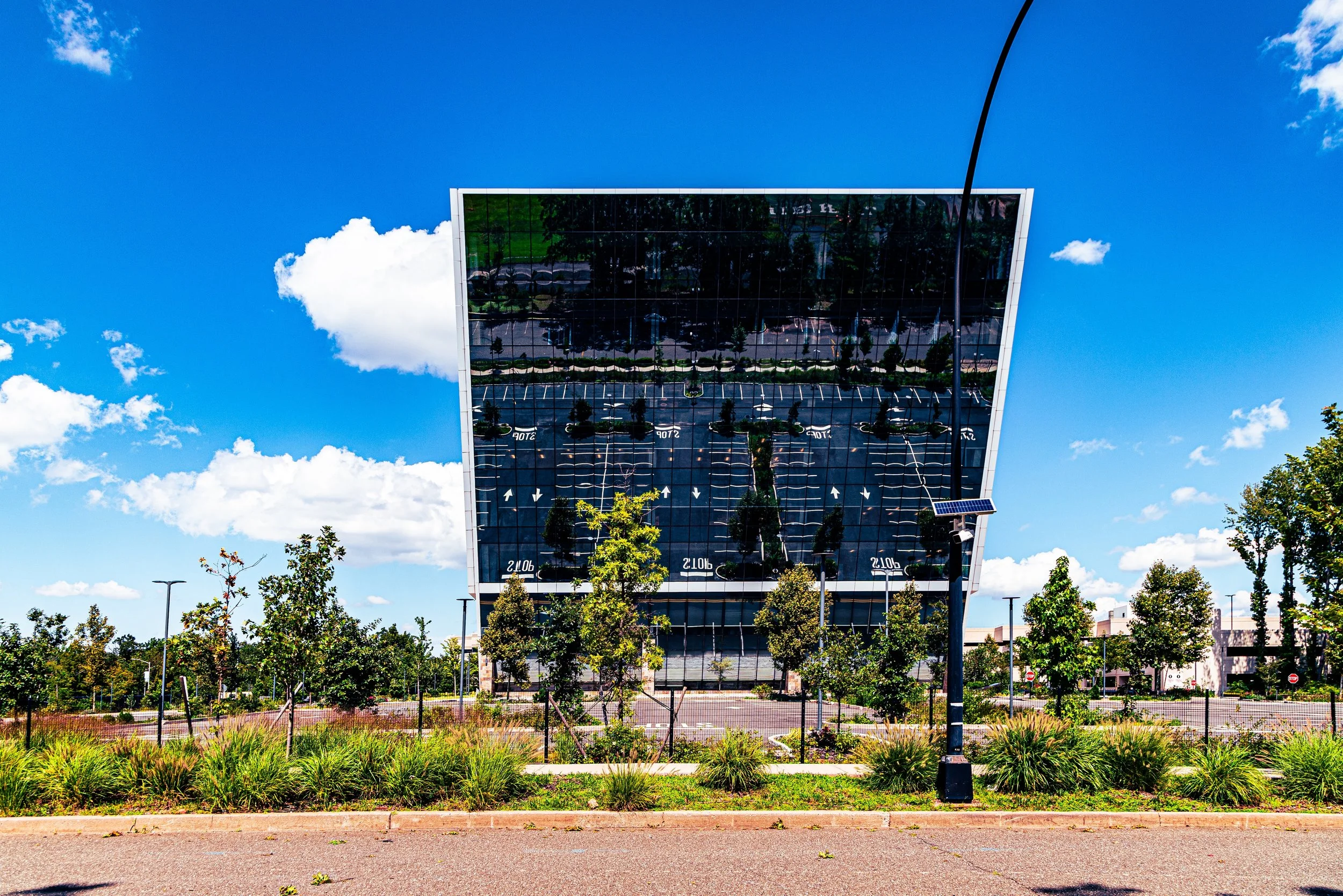 A modern building with a reflective glass facade, surrounded by trees and a parking lot, under a bright blue sky with scattered clouds.