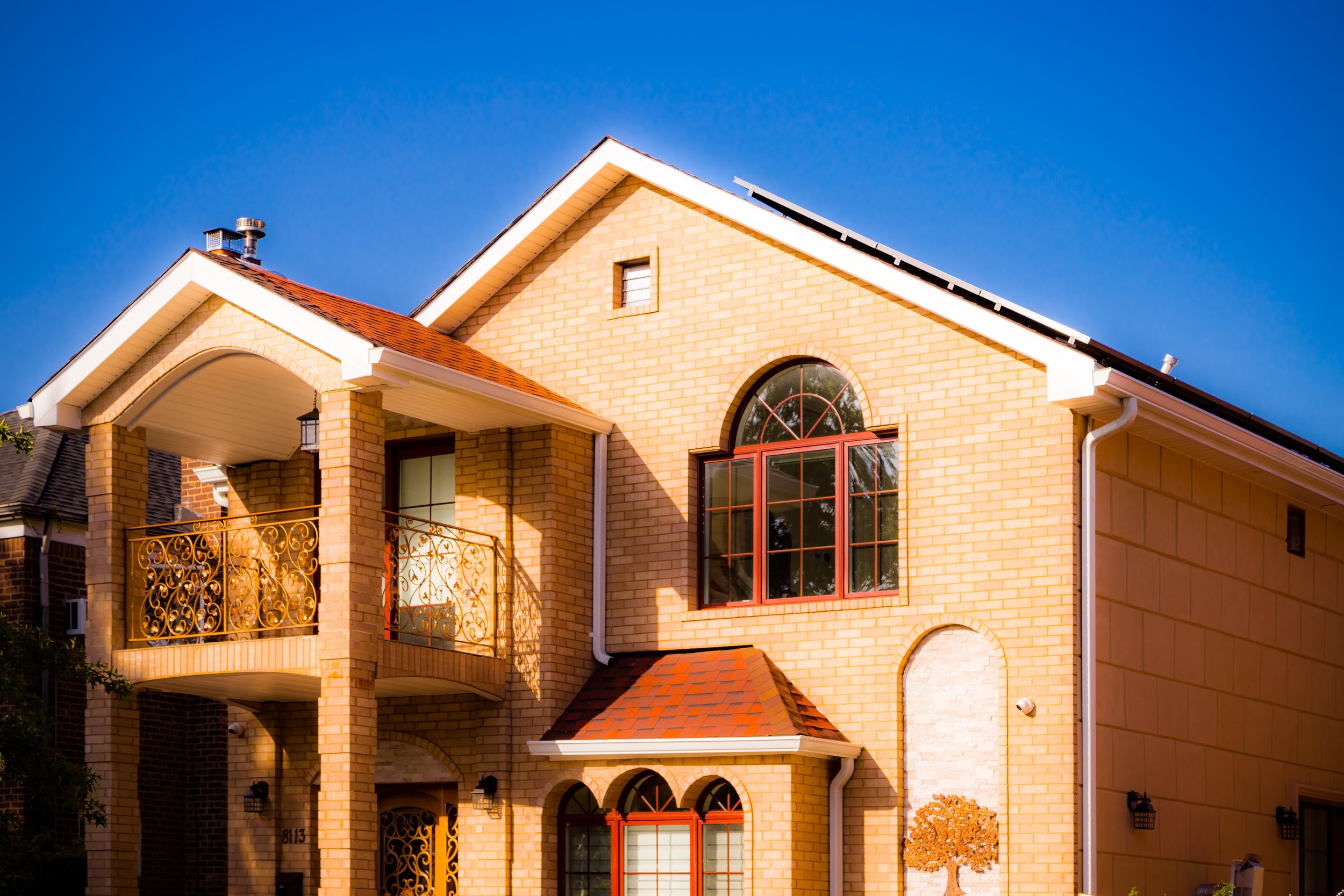 A yellow brick house with arched windows, a curved balcony with ornate metal railing, and a red tile roof, under a clear blue sky.