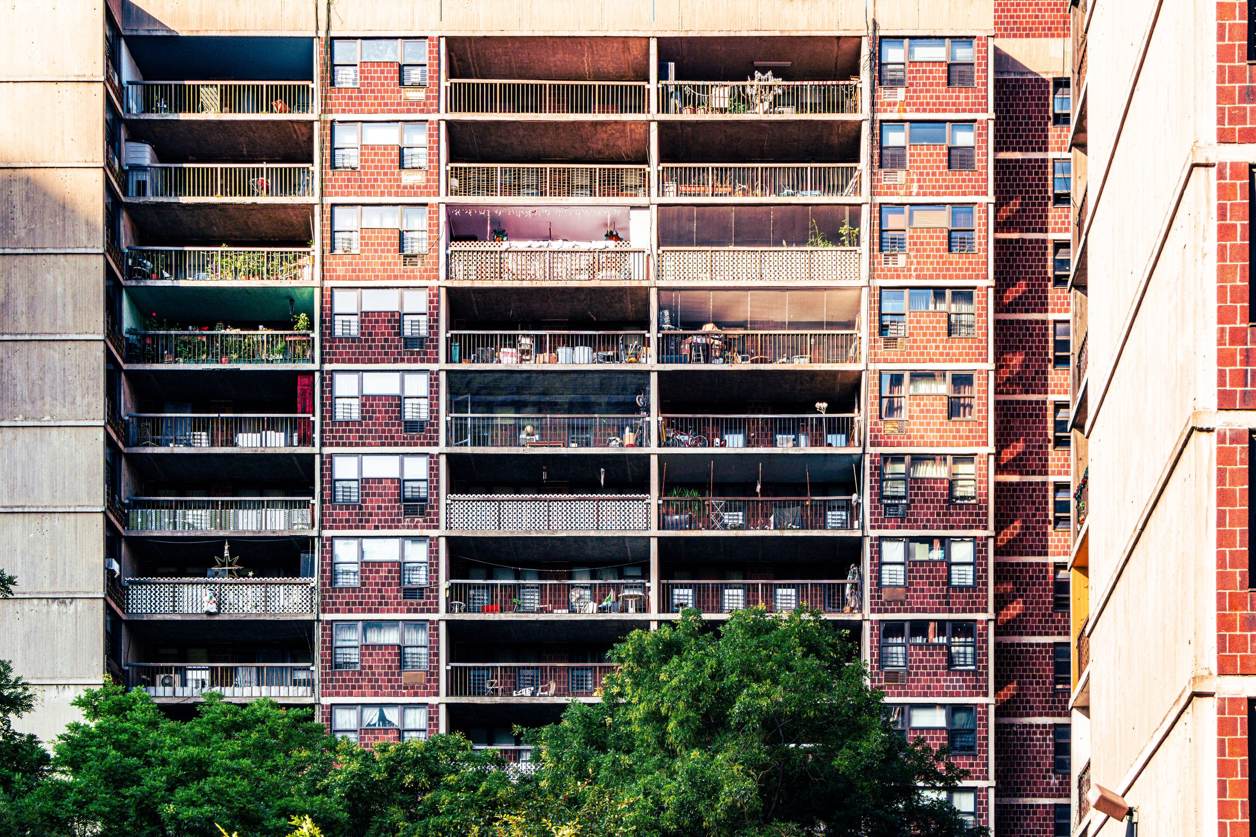 Exterior view of a multi-story apartment building with balconies, some with plants and outdoor furniture, and a large green tree in front