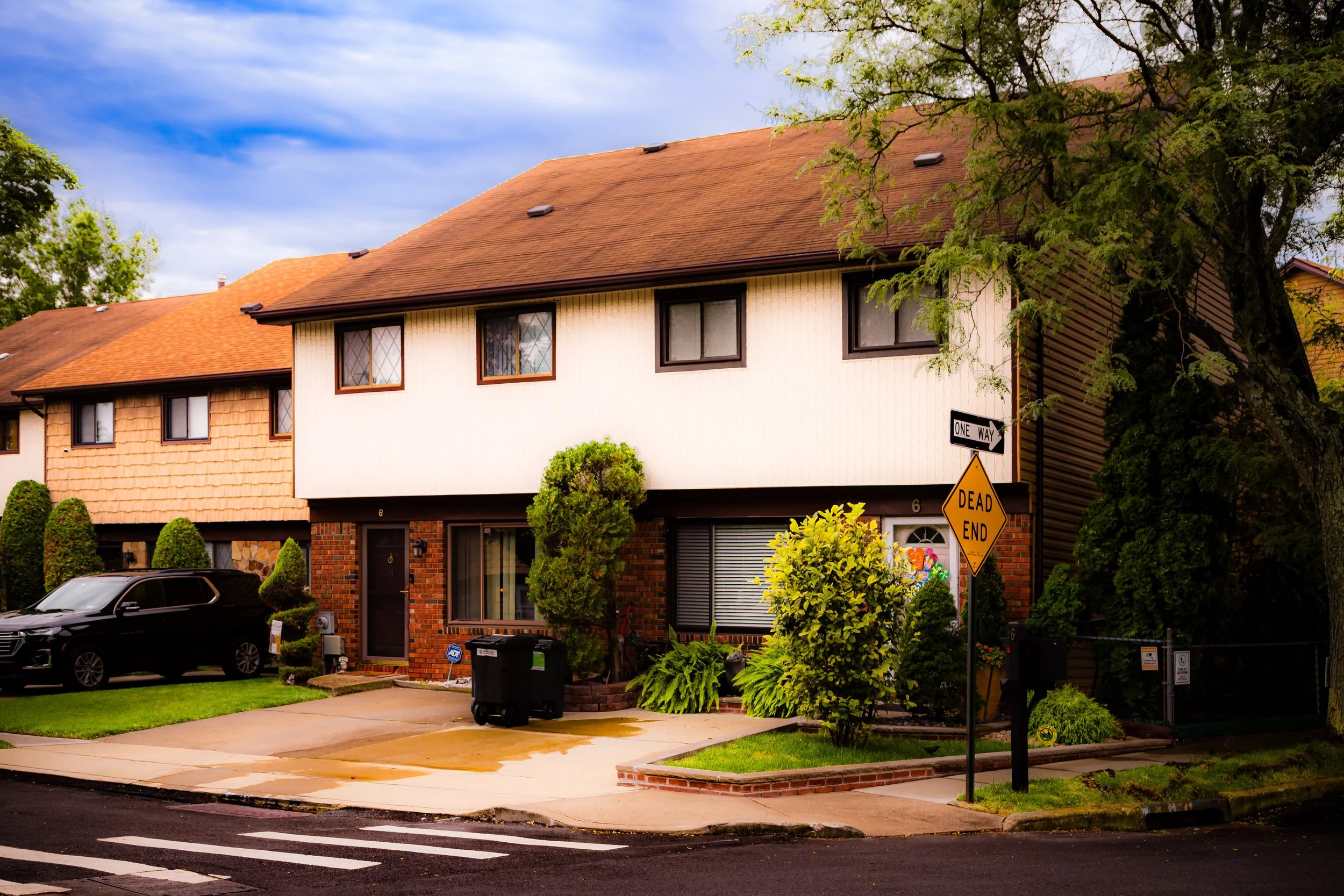 A two-story suburban house with a brick lower level and white upper level, with a brown roof, surrounded by greenery and parked car in the driveway.