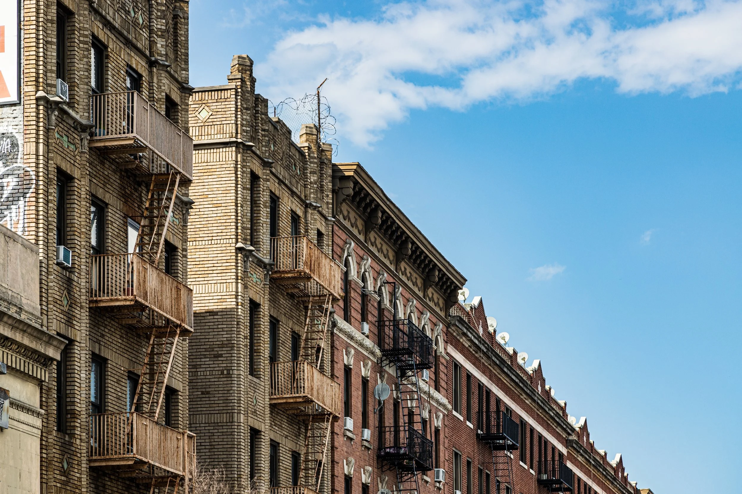 Red and beige brick apartment buildings with fire escapes and satellite dishes against a blue sky with some clouds.